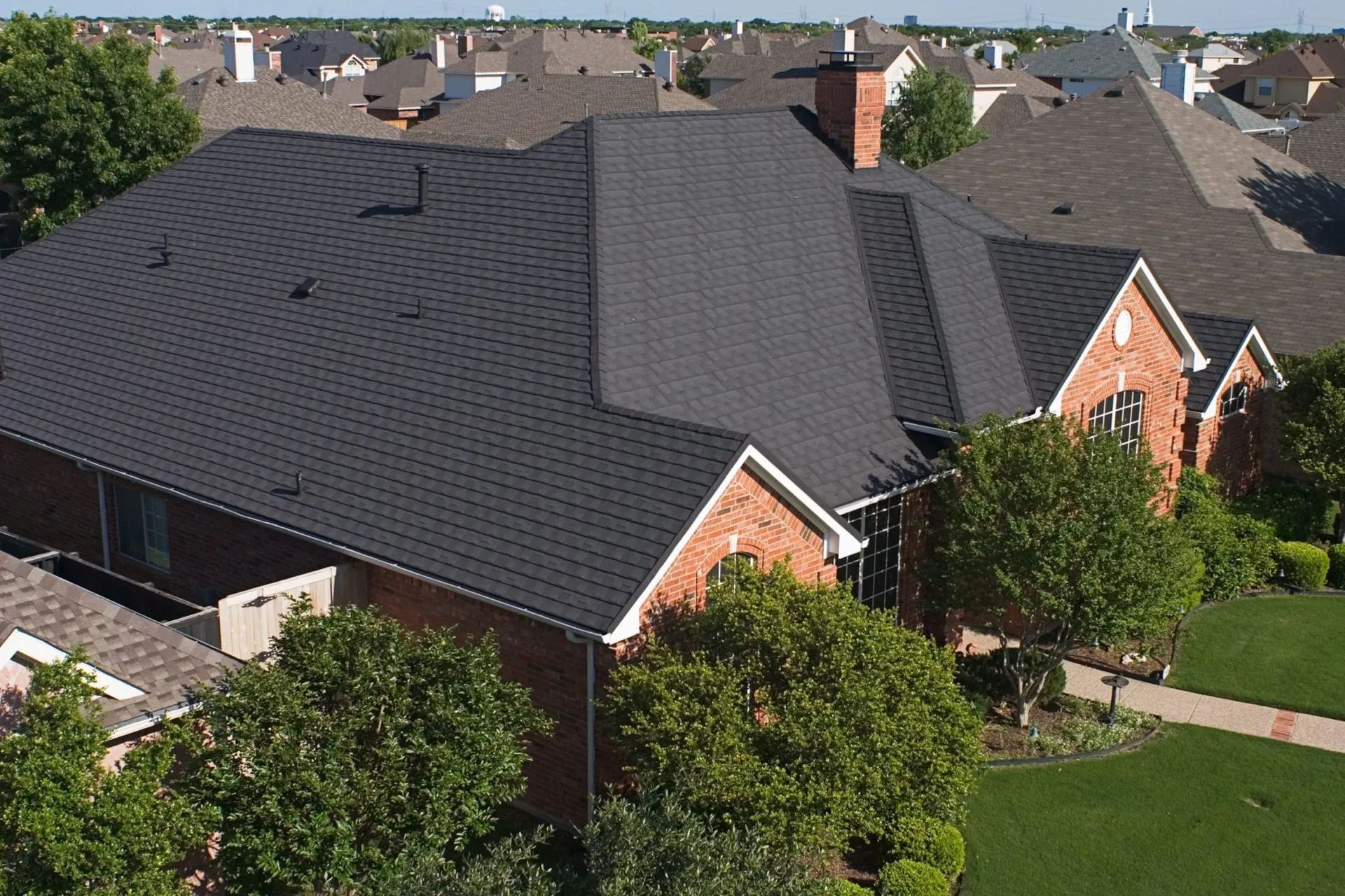 An aerial view of a brick house with a dark shingled roof in a suburban neighborhood.