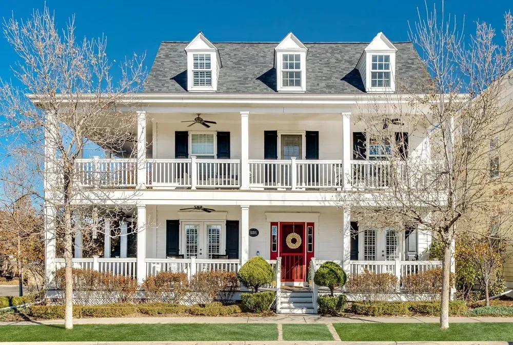 A white, two-story house with a full-width front porch on both levels, three roof dormers, and a bright red front door.