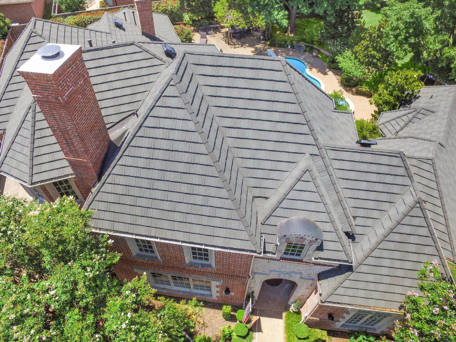Aerial view of a multi-gabled brick home with a gray shingled roof, surrounded by trees and a pool.
