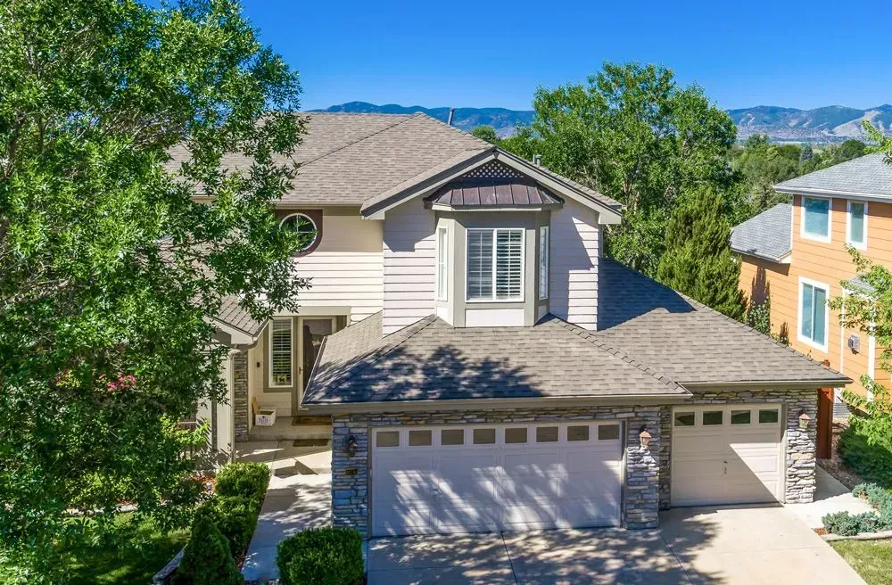A two-story tan suburban house with a gray roof and a two-car garage, set against a backdrop of trees and mountains.