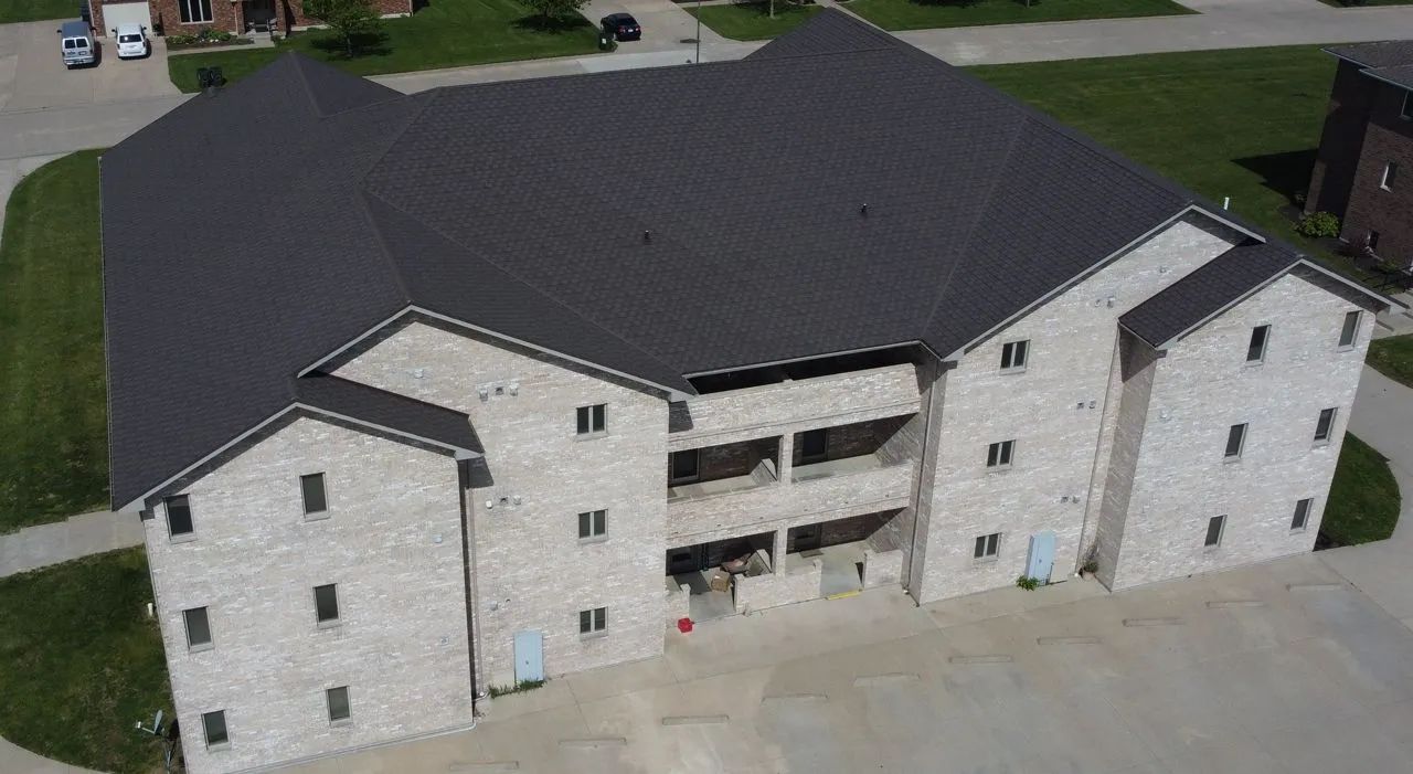 An aerial view of a three-story light-colored brick apartment building with a dark shingled roof and a parking lot.