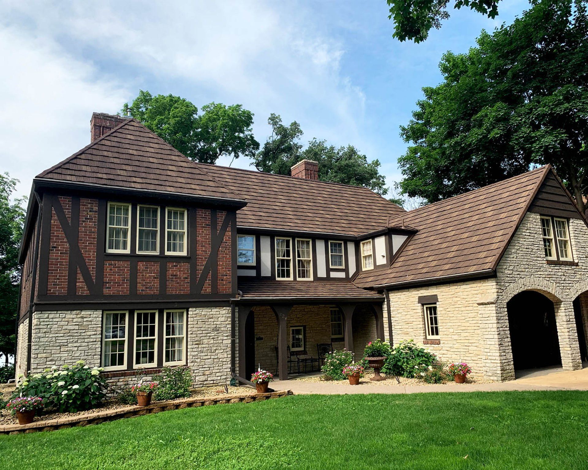 A two-story Tudor-style home with timber-framed brick, stone masonry, a brown shingled roof, and a grassy front lawn.