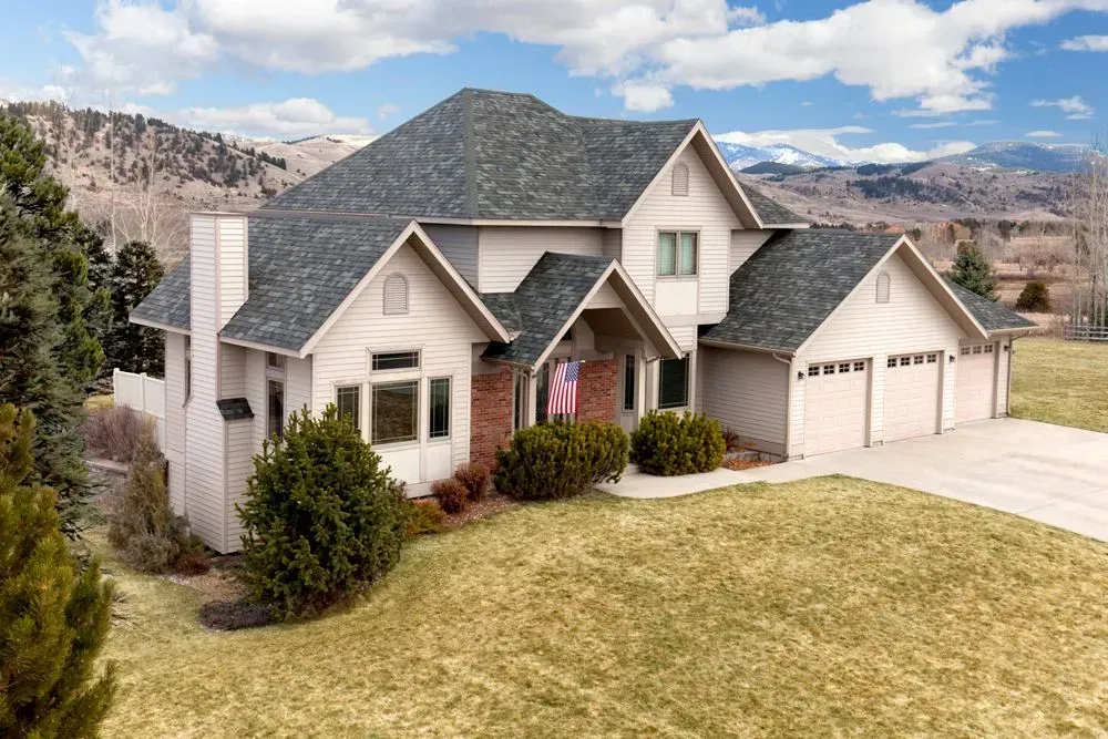 A two-story, light beige house with a dark gray shingled roof and a multi-car garage, set against a mountainous landscape.