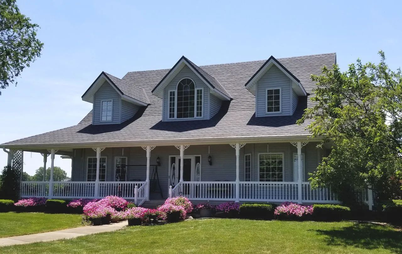 A grey two-story house with a wraparound porch, three gabled dormers, and pink bushes in the front yard.