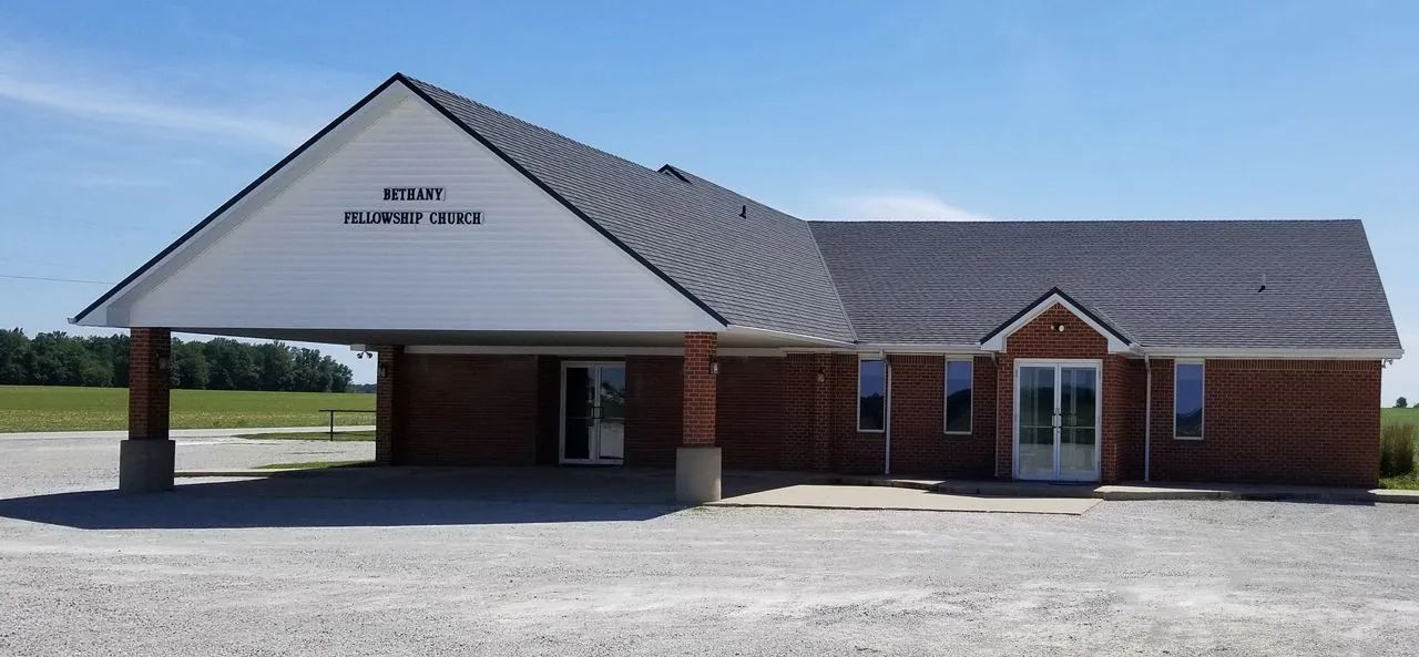 A modern red brick building with a large white covered entrance, surrounded by a gravel parking lot under a blue sky.
