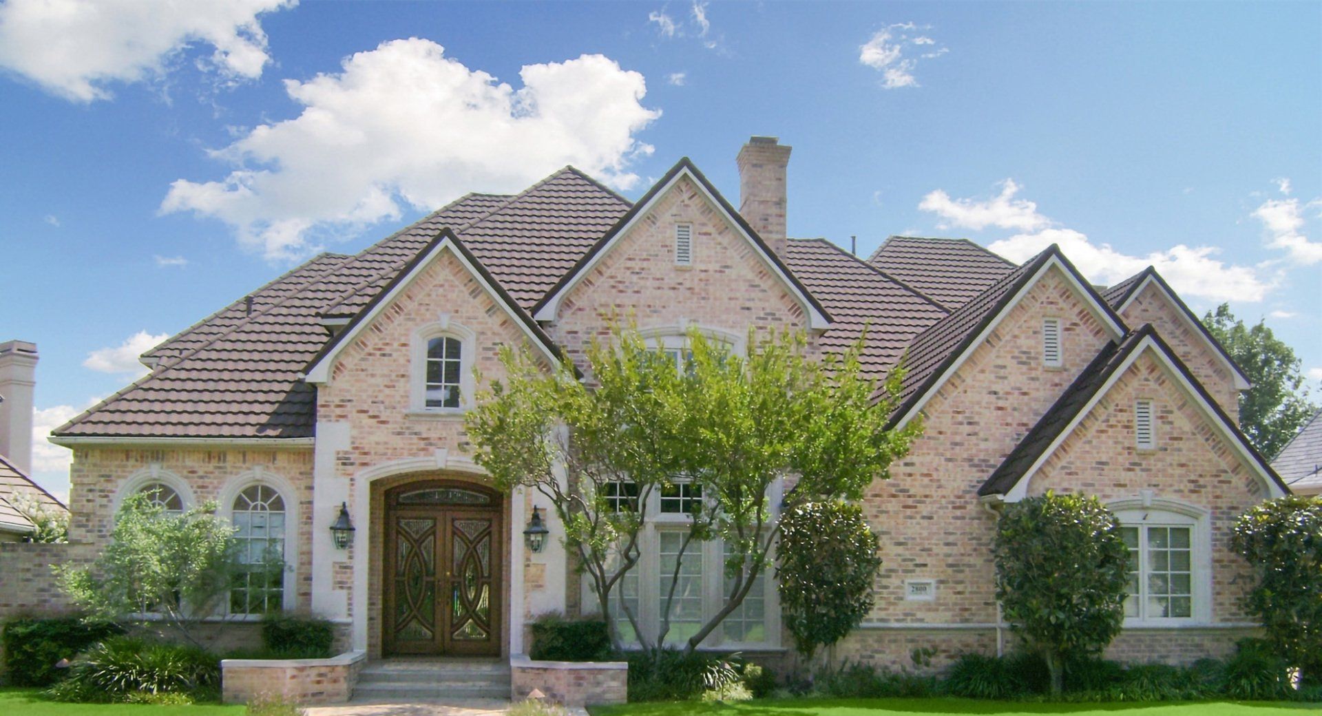 A two-story house with light brick siding, a brown roof, large front doors, and manicured landscaping under a blue sky.