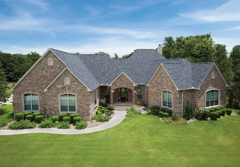 A large, single-story stone house with a multi-gabled dark roof, surrounded by a green lawn and mature trees.