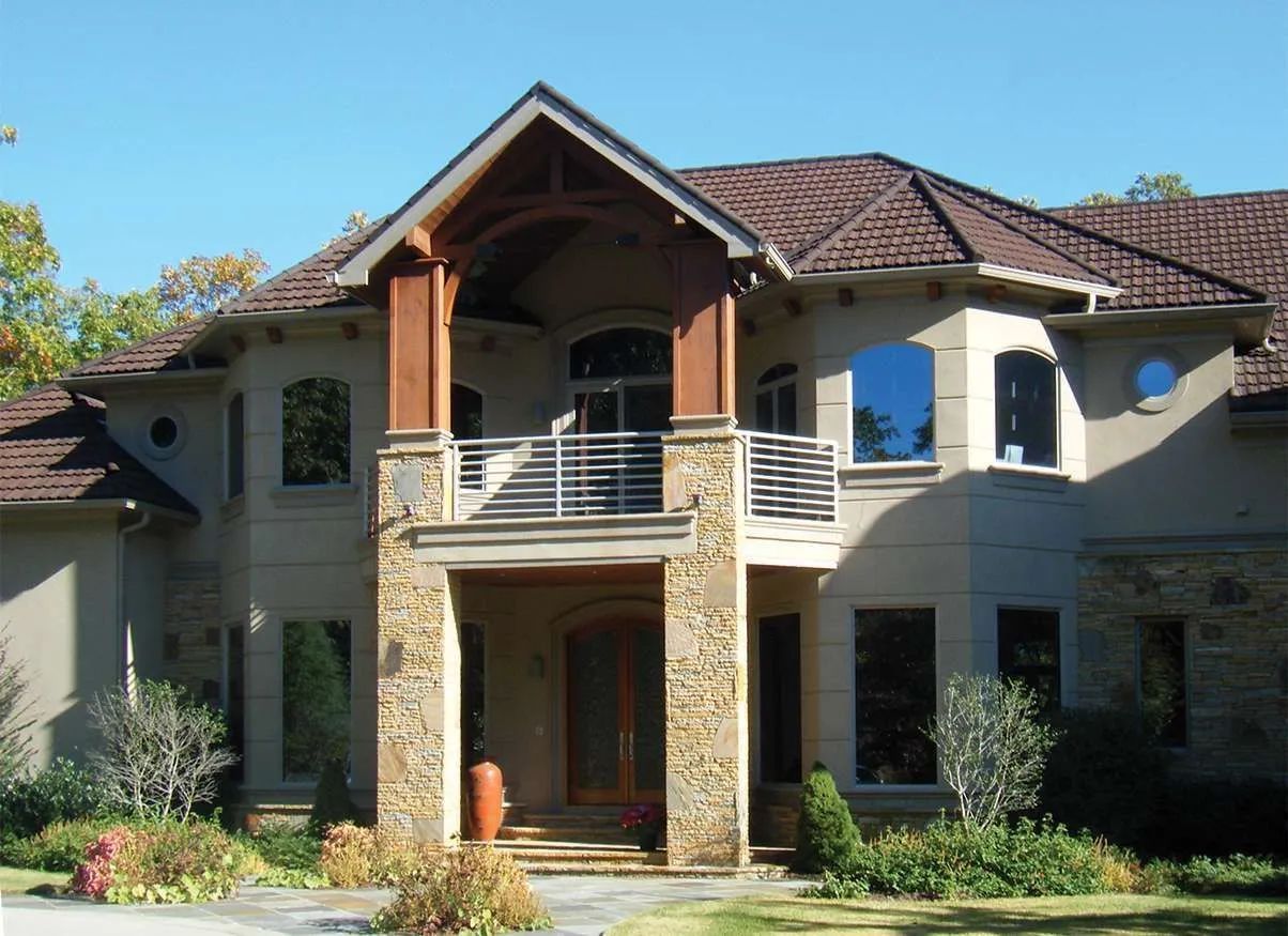 A large, two-story stone and stucco home featuring a covered balcony, dark roof tiles, and multiple windows.