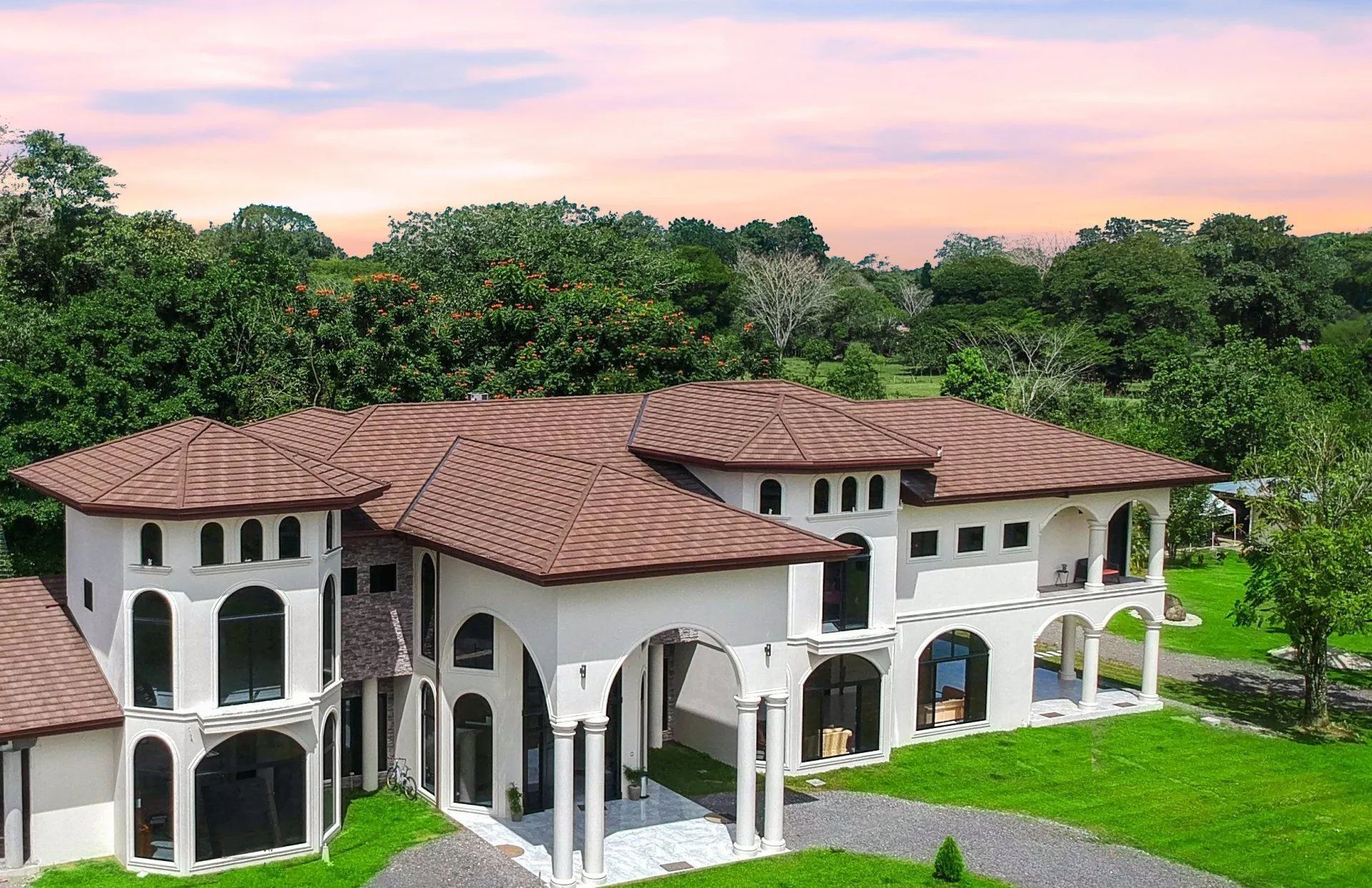 A large, two-story white Mediterranean-style villa with a brown tile roof, surrounded by green trees and a lawn.