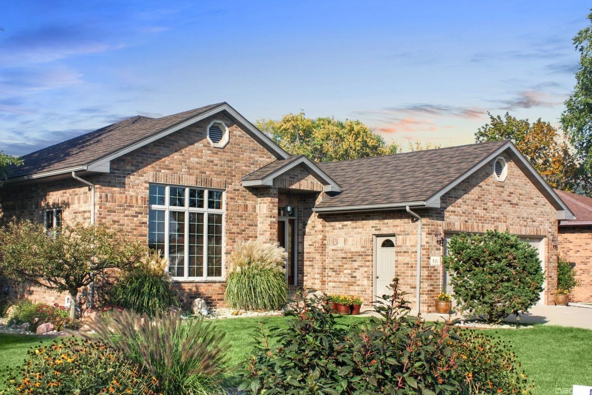A single-story, tan brick house with a brown shingled roof, large windows, and a landscaped front yard under a blue sky.
