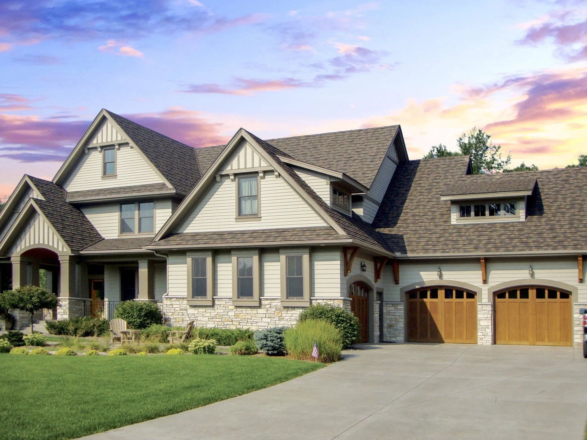 A large, two-story craftsman-style house with beige siding, stone accents, and a three-car garage at sunset.