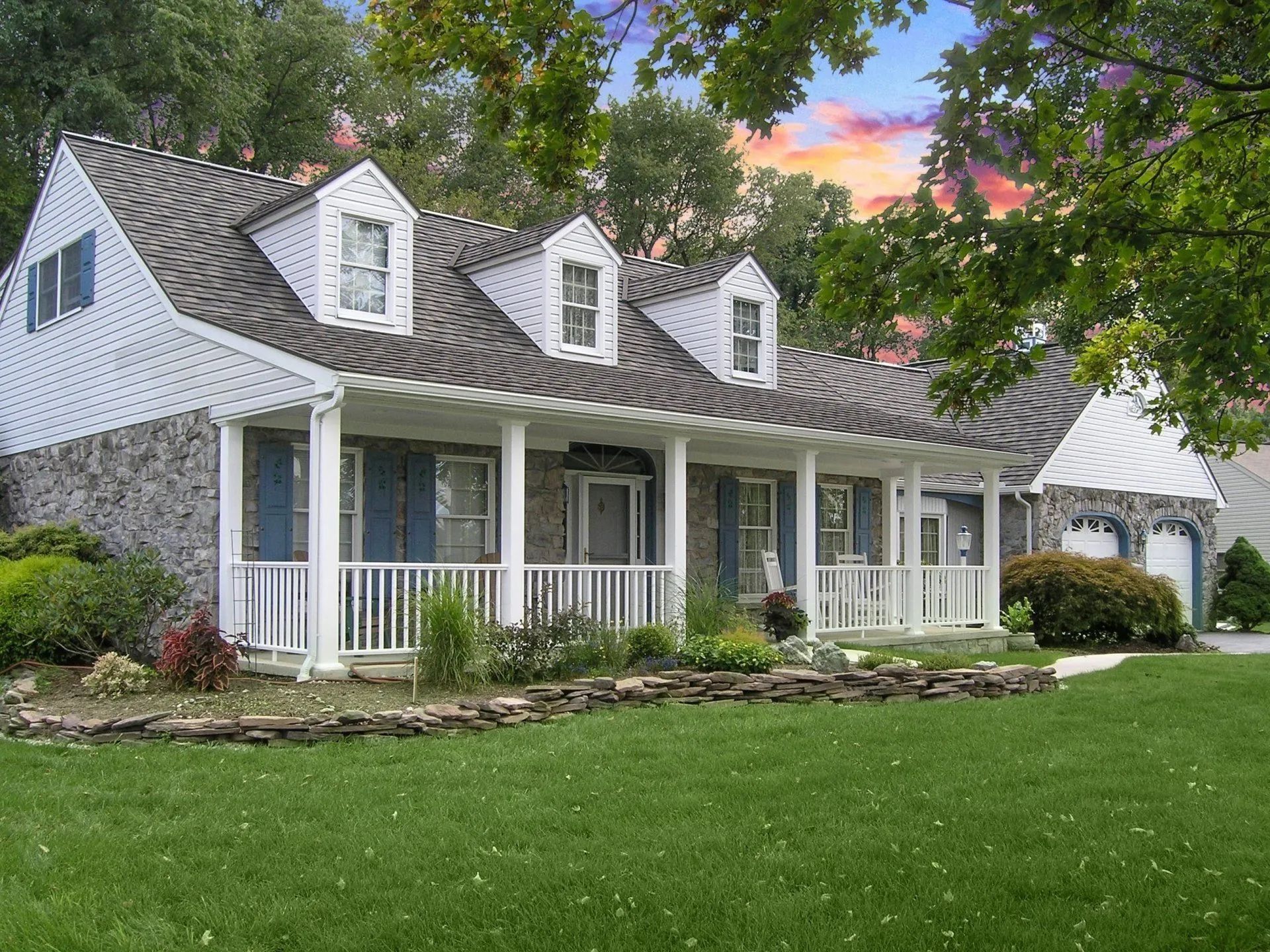 A stone-and-siding farmhouse with three dormers, a full-length covered porch, and a manicured lawn under a sunset sky.