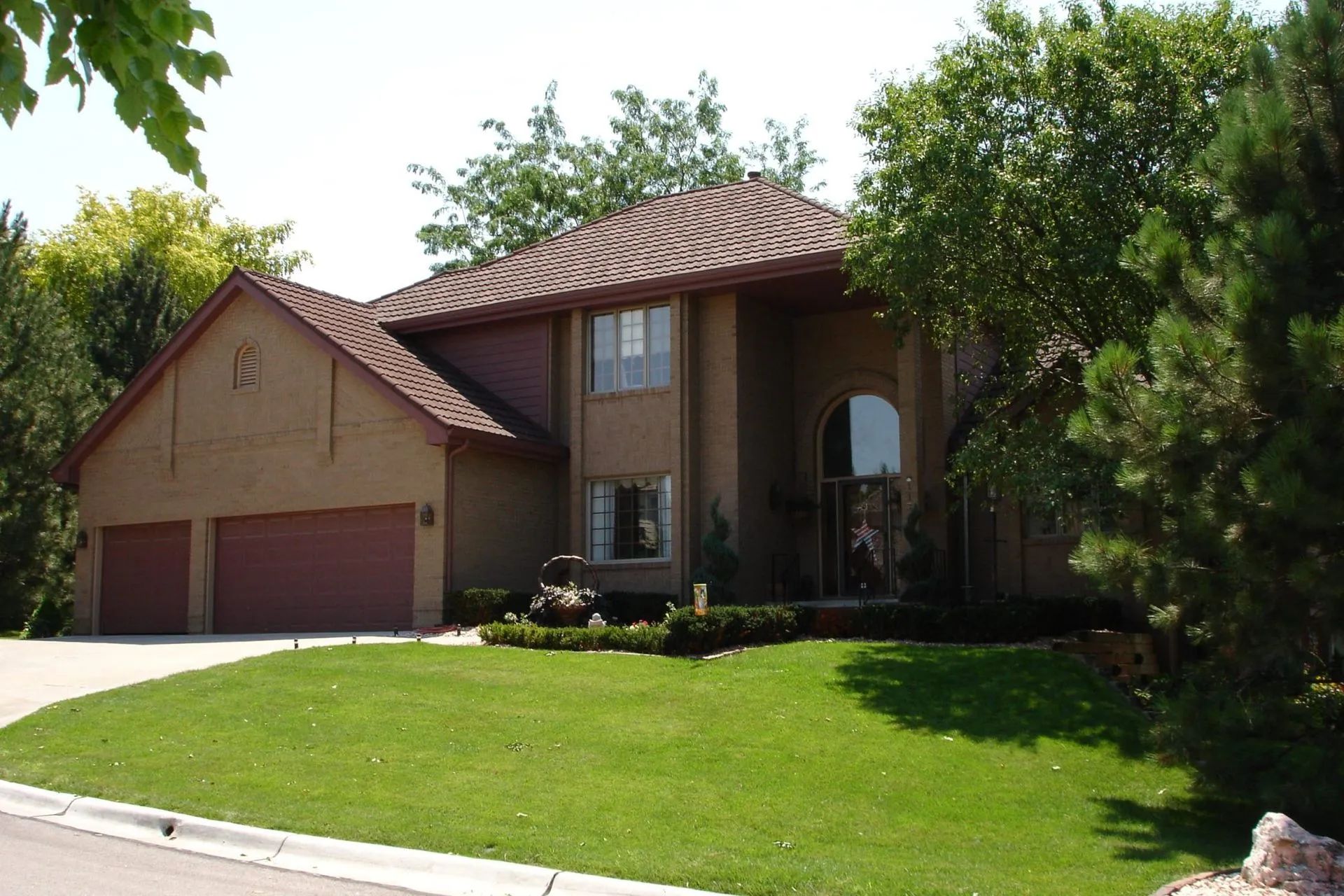 A tan two-story house with a brown roof and attached garage, set behind a lush green lawn and surrounded by trees.