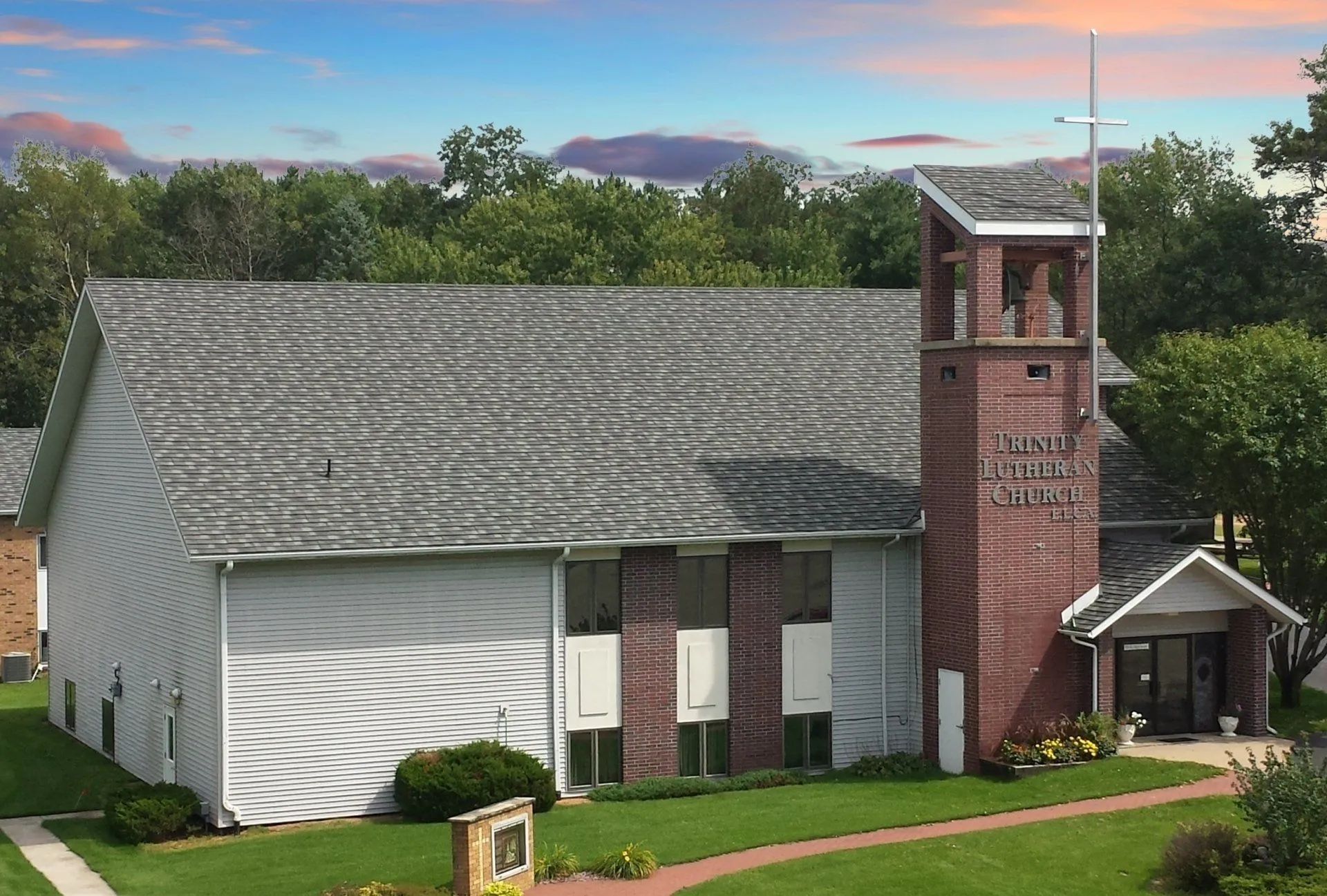 A church with a grey shingled roof, light siding, a tall brick bell tower, and a cross against a sky with soft clouds.