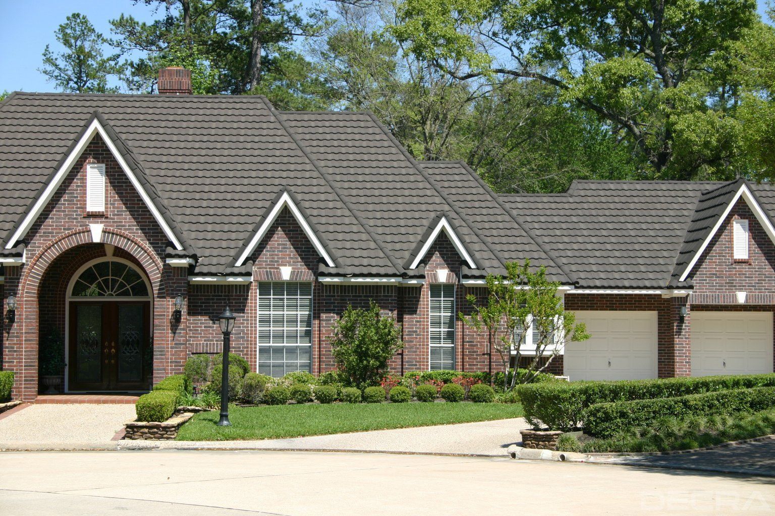 A one-story brick house with a dark gabled roof, arched entryway, and attached three-car garage, surrounded by landscaping.