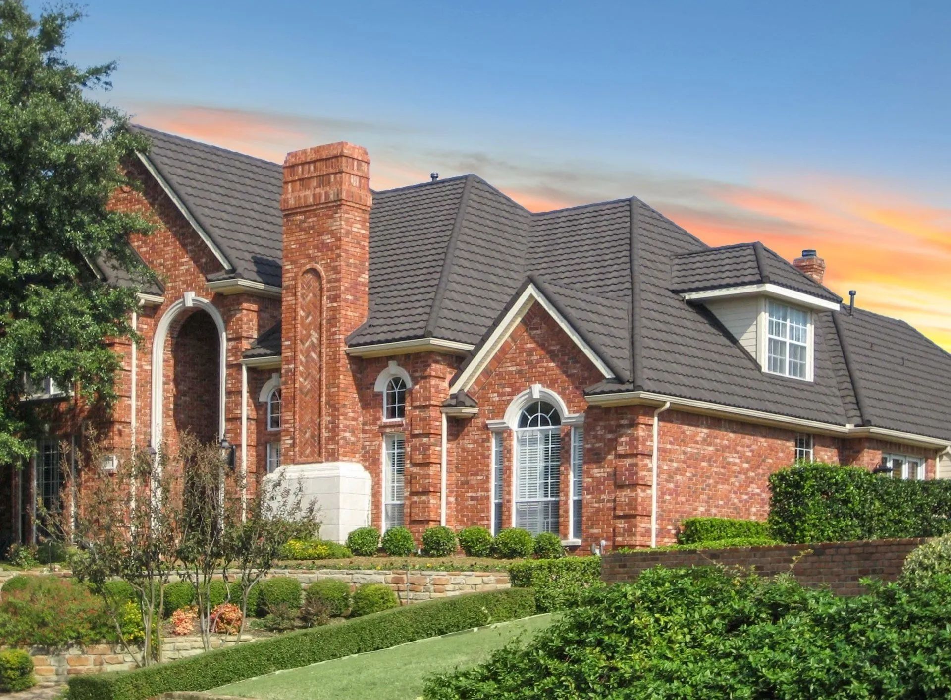 A large, multi-story red brick house with a dark gabled roof, tall chimney, and landscaped garden under a sunset sky.