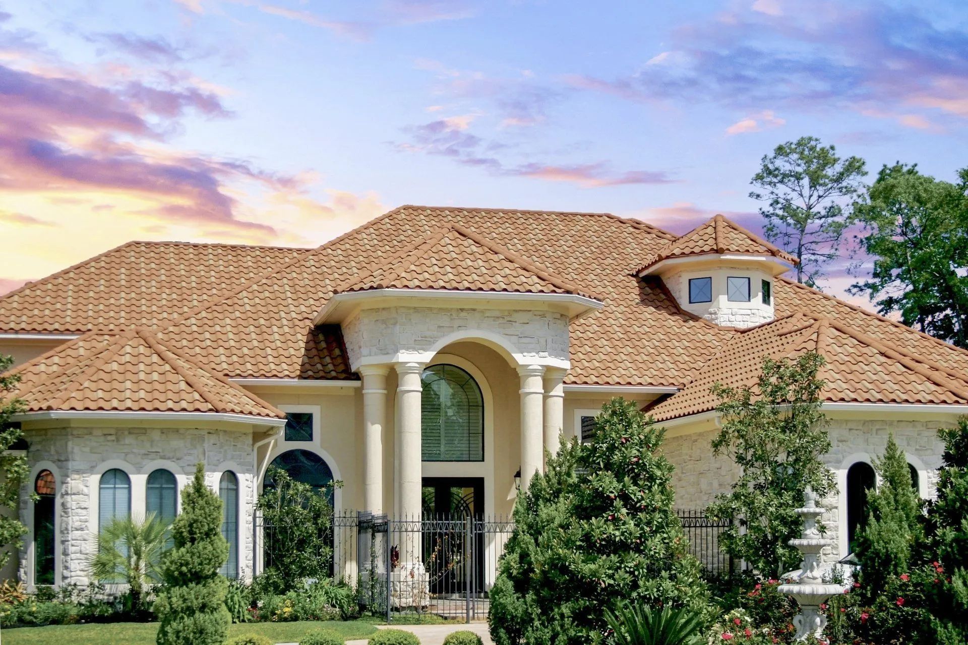A tan stone house with a multi-gabled terracotta tile roof, columns at the front entrance, and lush green landscaping.