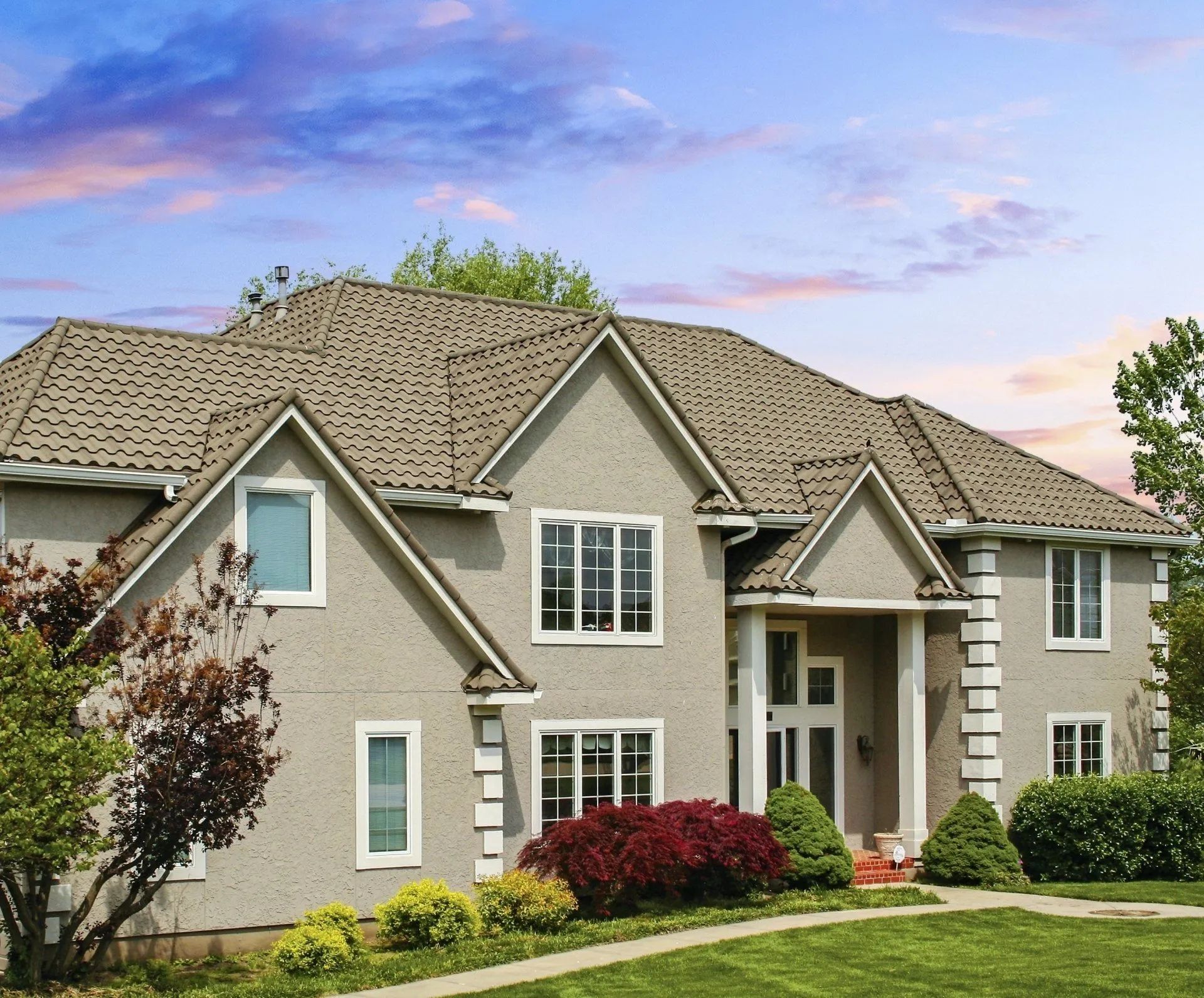 A two-story stucco suburban house with a brown shingled roof, light siding, white trim, and a neatly landscaped front yard.