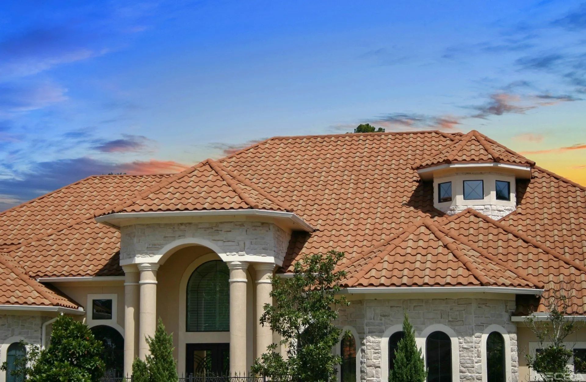 A sprawling luxury home with a textured, reddish-brown tiled roof and beige stone exterior under a vibrant sunset sky.