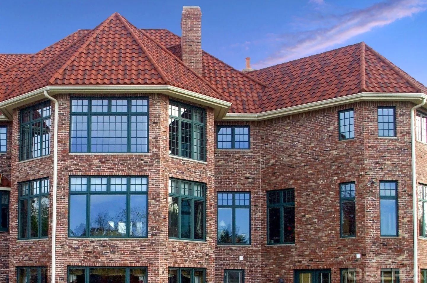 A large, two-story brick house with a red tile roof and dark-framed windows under a blue sky.