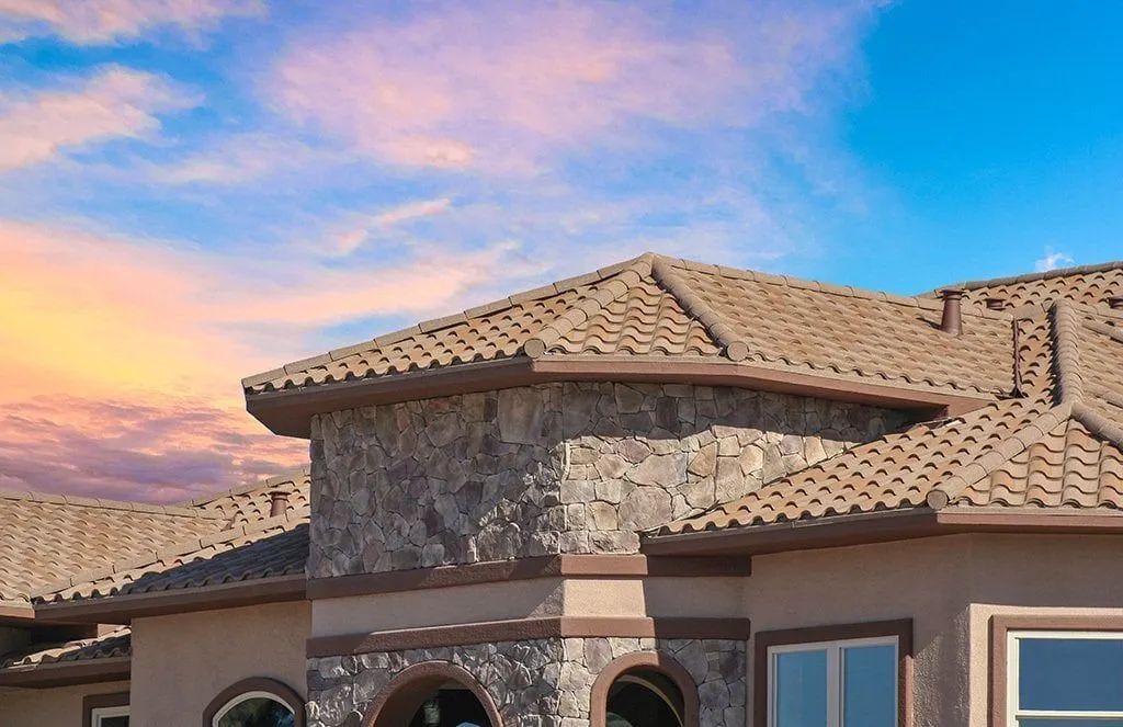 The upper exterior of a stucco home with stone siding and a brown tile roof under a blue sky with pink sunset clouds.