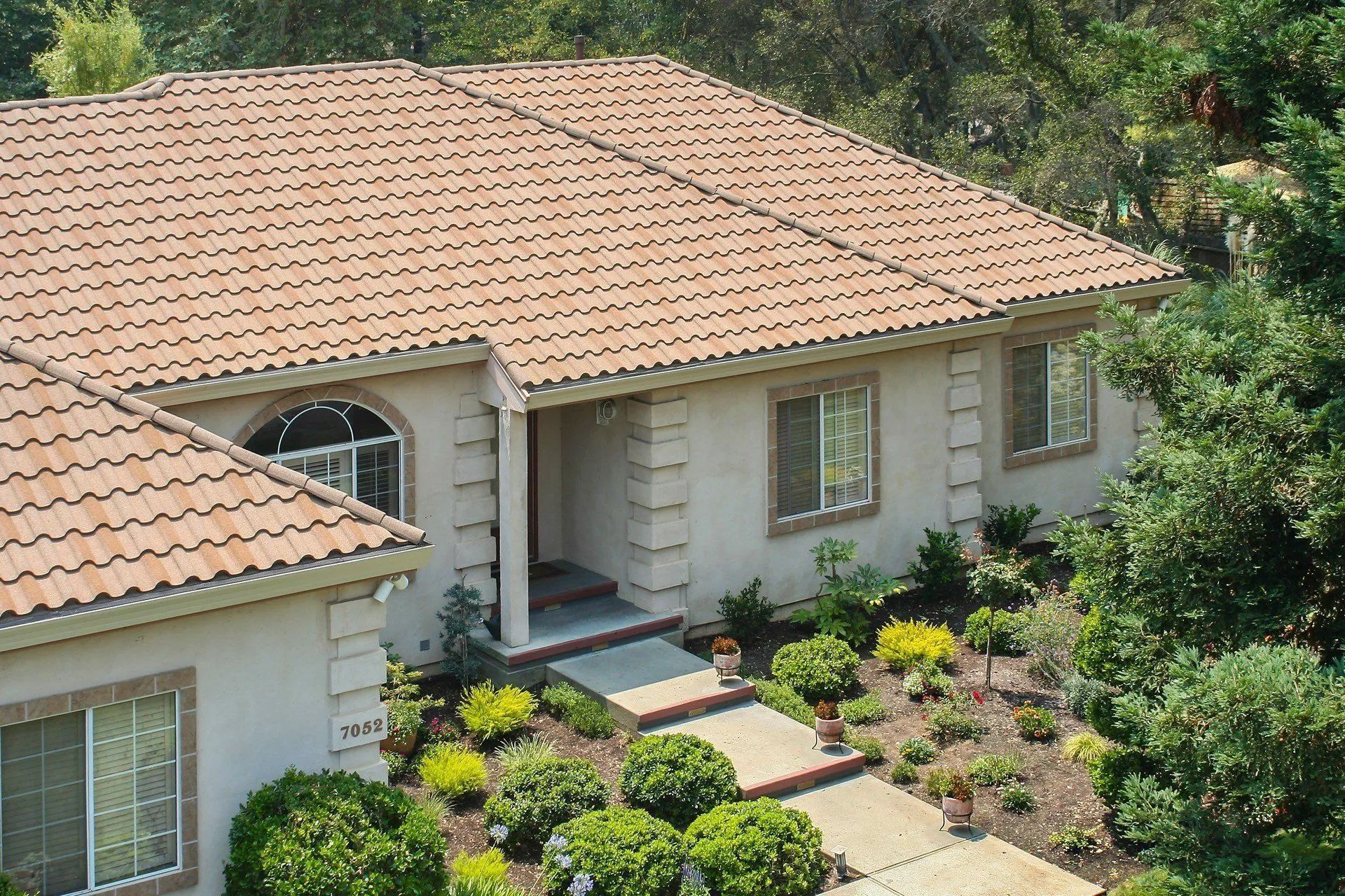 A suburban house with a tan, arched-tile roof, stucco exterior, and a landscaped front yard with green bushes.