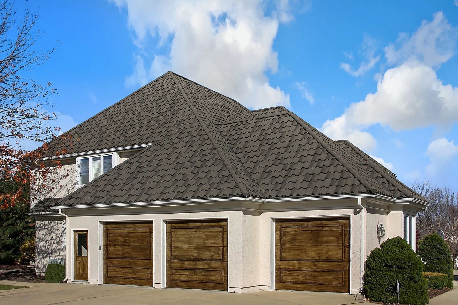 A multi-level home with a beige stucco exterior, a dark shingled roof, and three dark wooden garage doors under a blue sky.