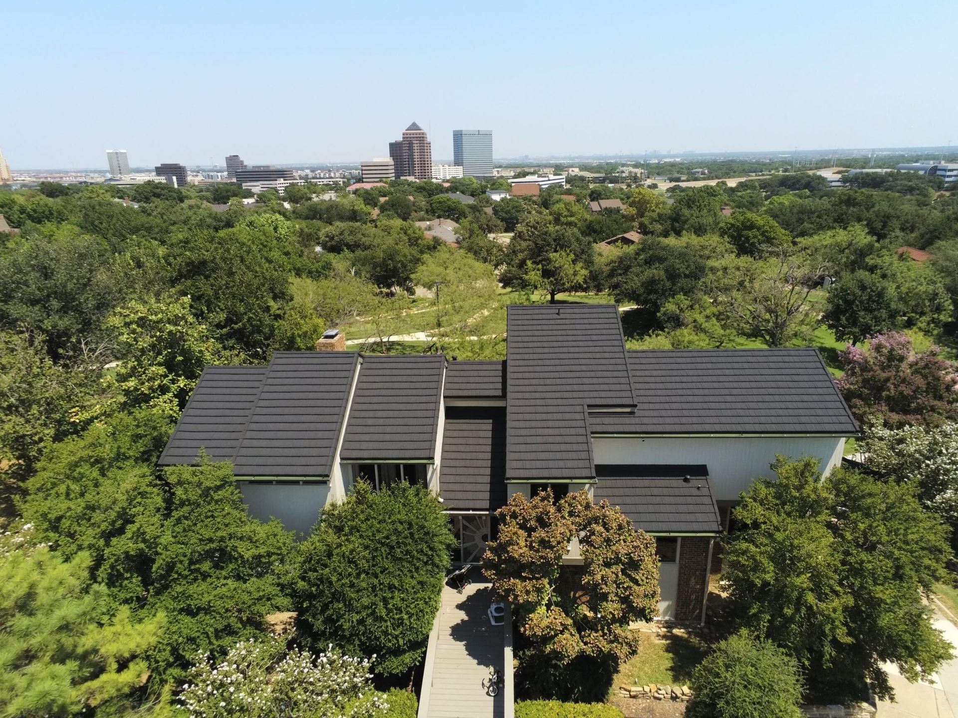 An aerial view of a modern house with a multi-layered, dark roof nestled in trees, with a city skyline in the distance.
