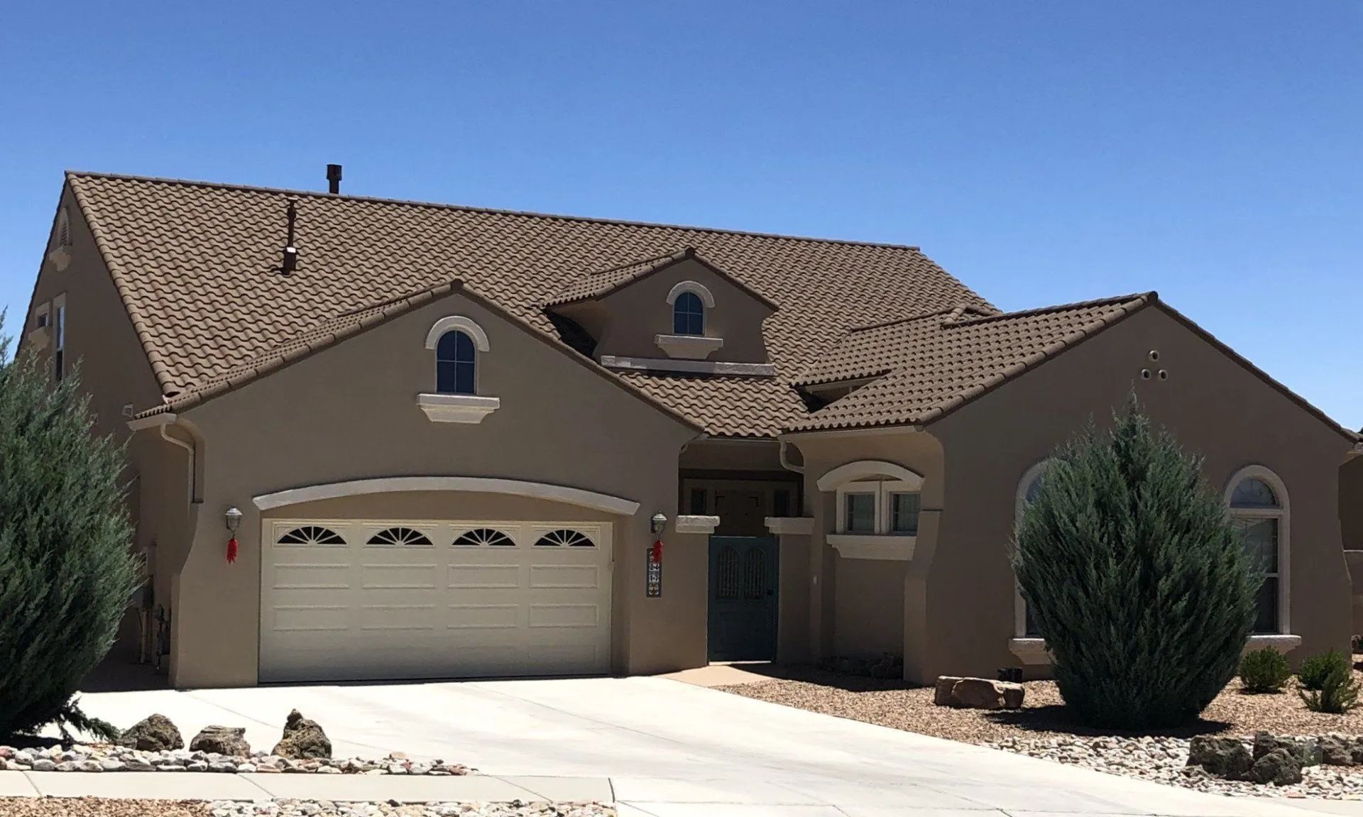 A tan, one-story house with a brown shingled roof, arched garage and window accents, and a concrete driveway.