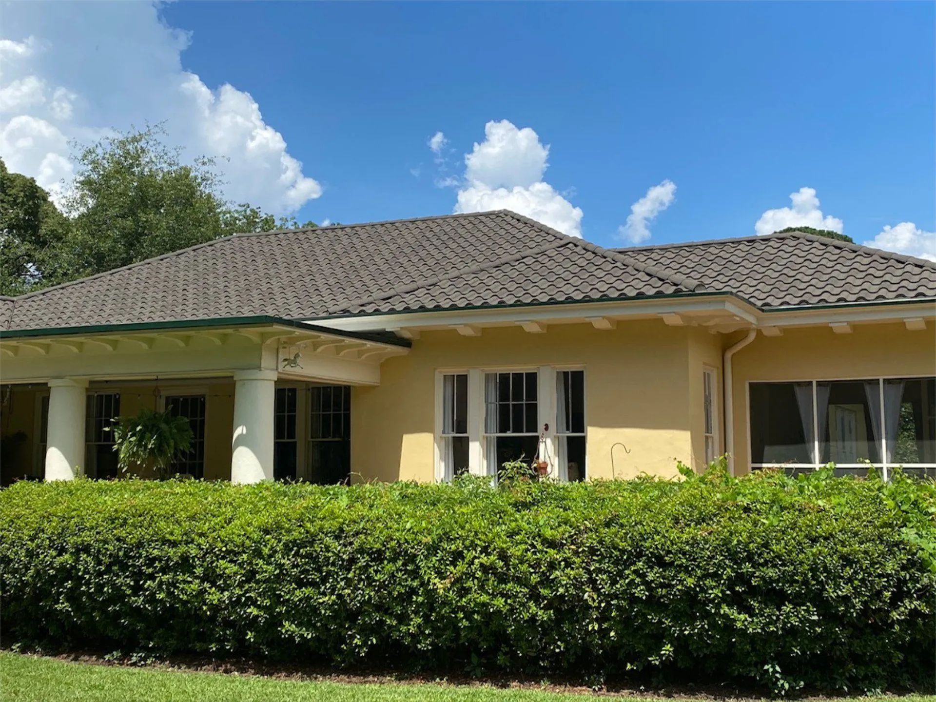 A single-story, light yellow stucco house with a dark tiled roof and a front porch, fronted by a large, manicured hedge.