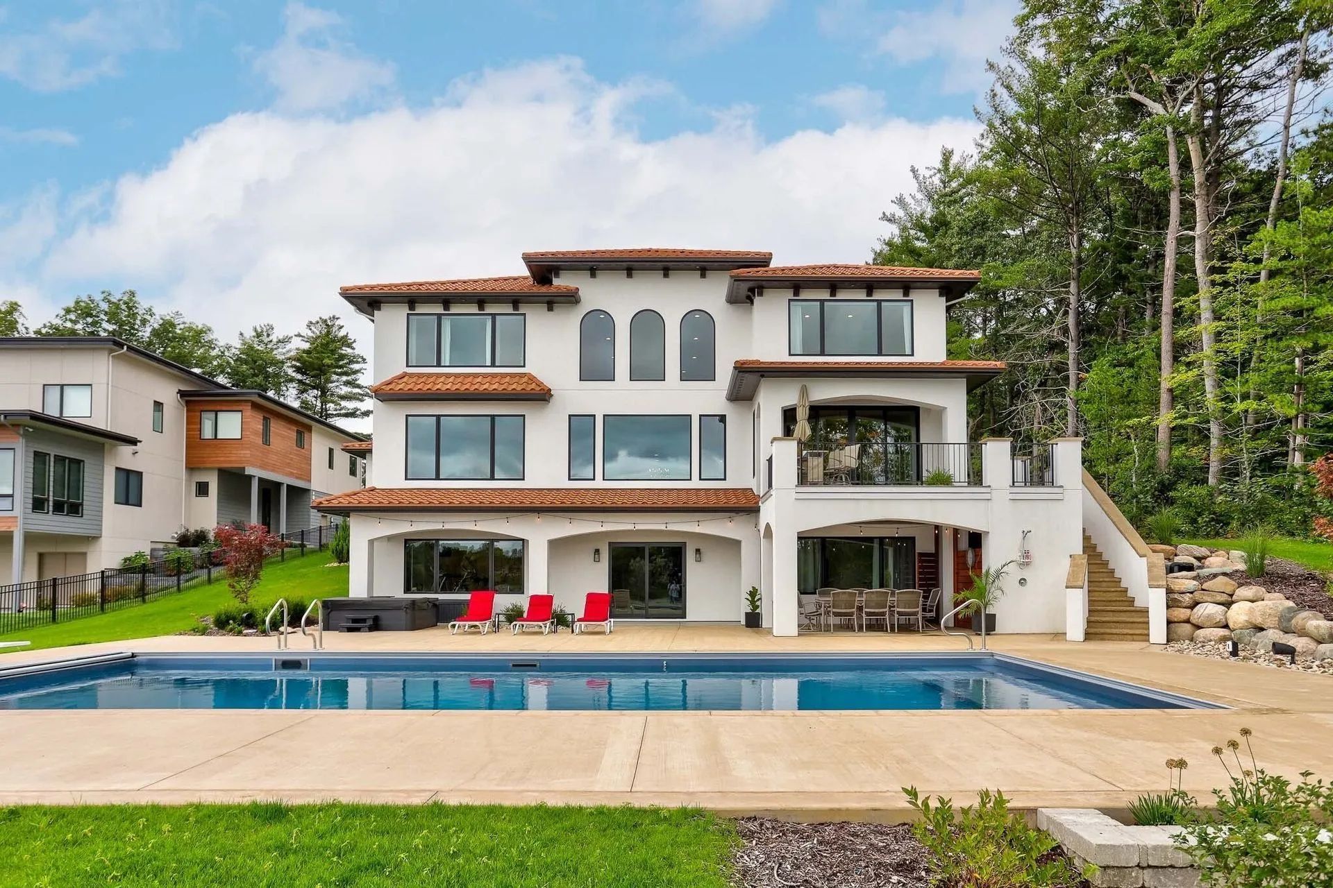 A multi-story villa with white stucco walls, red tile roofs, and a backyard swimming pool surrounded by a patio and trees.