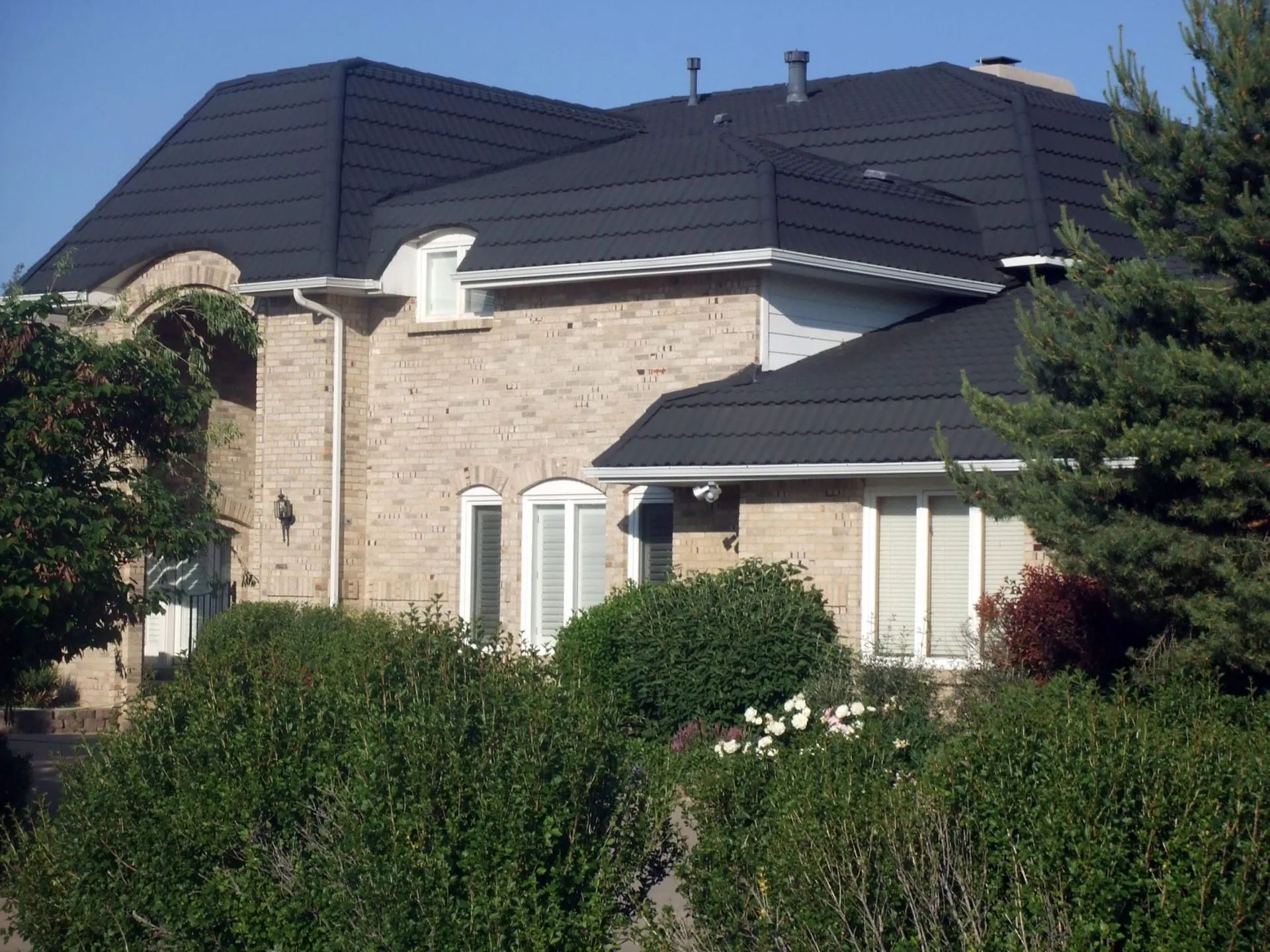 A two-story brick house with a dark, layered roof, surrounded by lush green trees and shrubs under a clear blue sky.