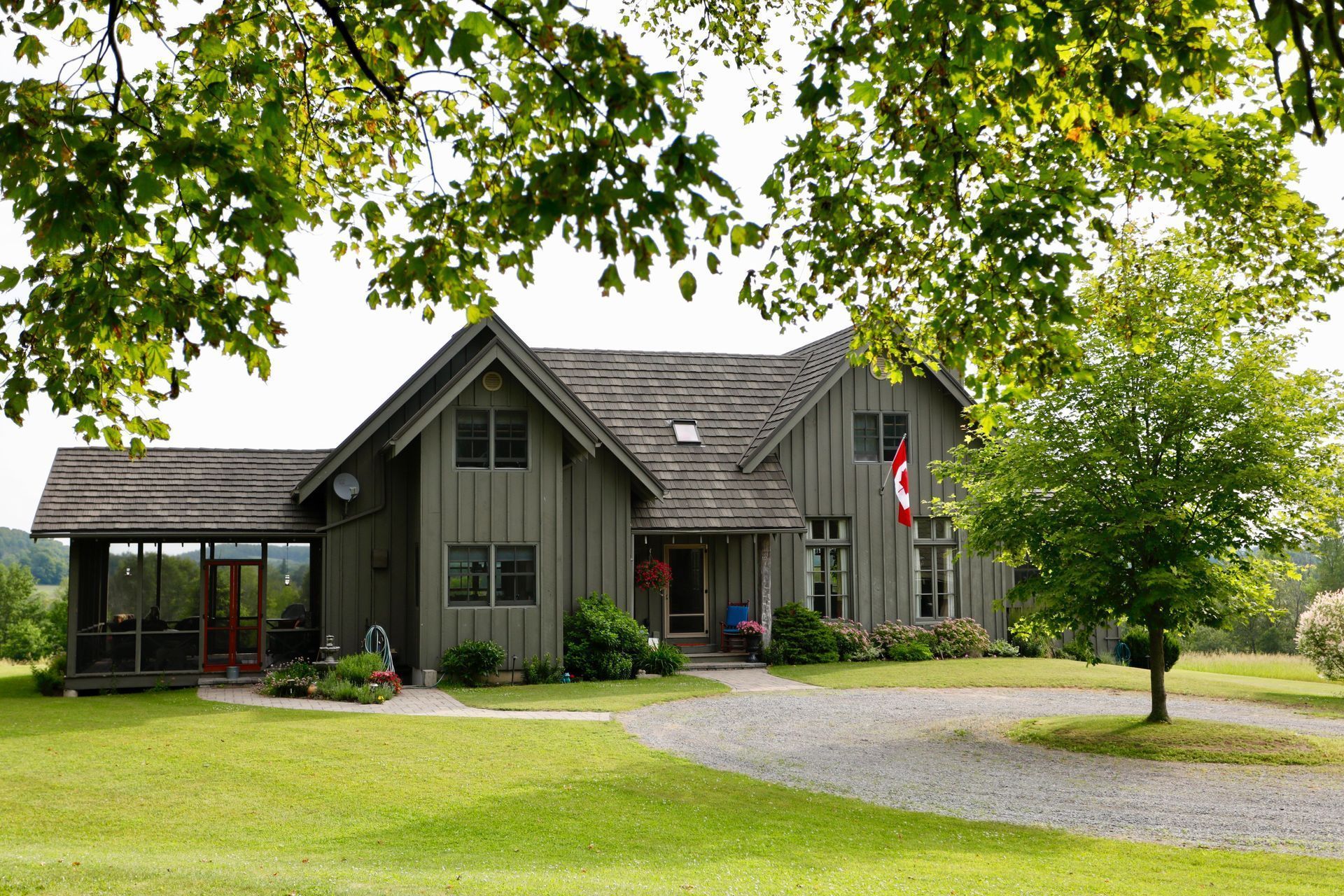 A rustic gray farmhouse with vertical wood siding and a covered porch, set on a lush lawn with a gravel driveway.