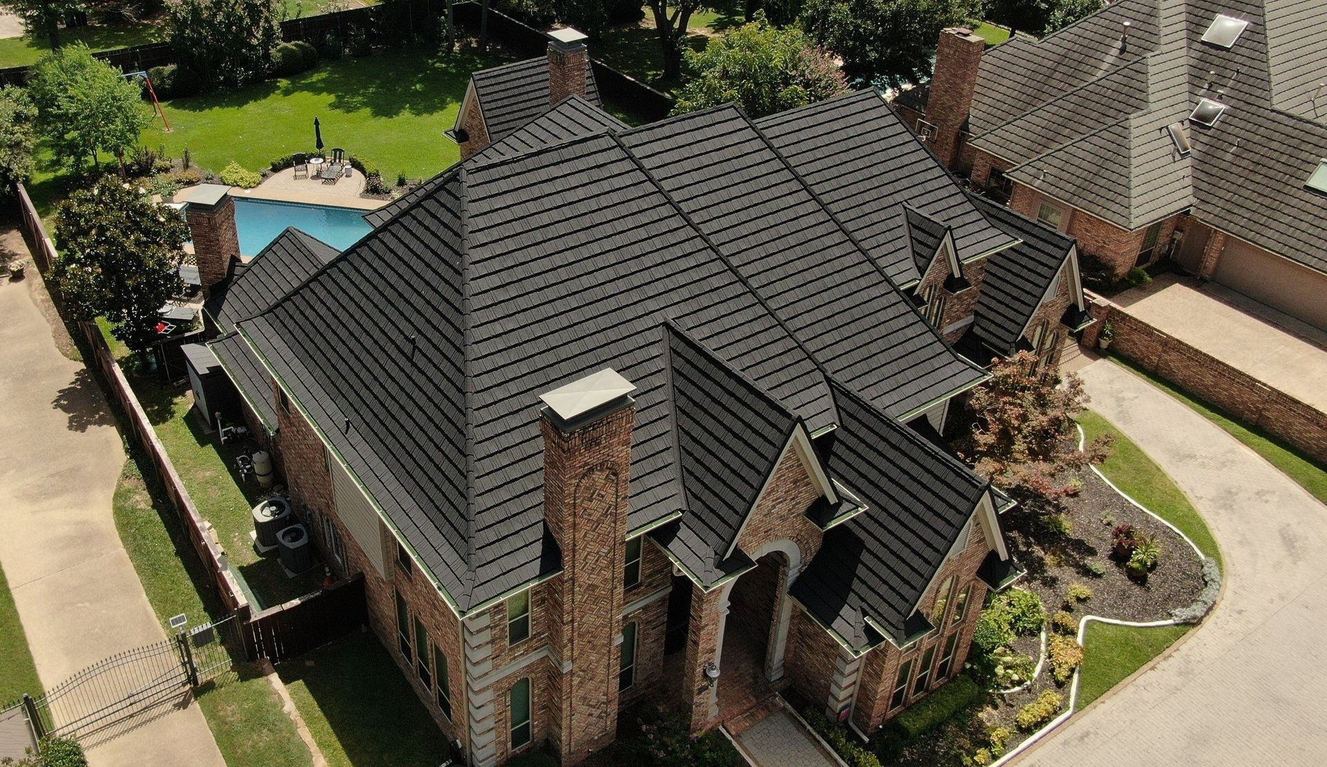 High-angle aerial view of a large brick house with a dark, textured gabled roof, driveway, and backyard pool.