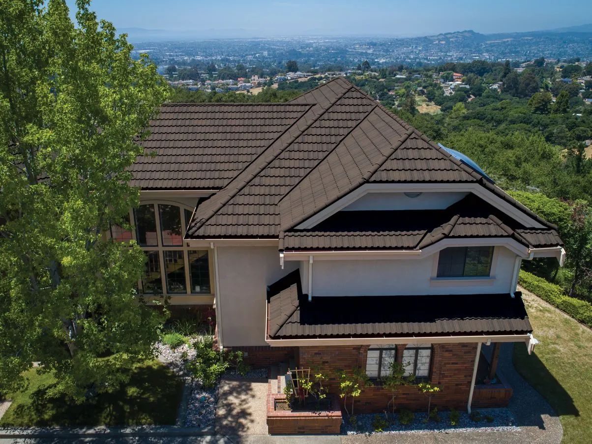 Aerial view of a two-story house with a brown metal roof, surrounded by trees, overlooking a distant suburban valley.