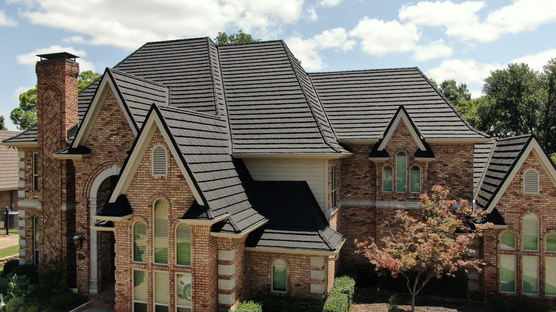 A two-story, multi-gabled brick home with a dark shingled roof, arched windows, and an exterior chimney on a sunny day.
