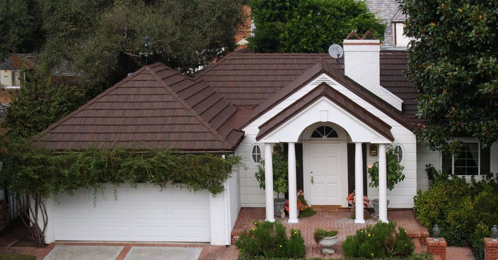 A white house with a dark brown tiled roof, a pillared entryway, and a garage, surrounded by green trees and shrubs.