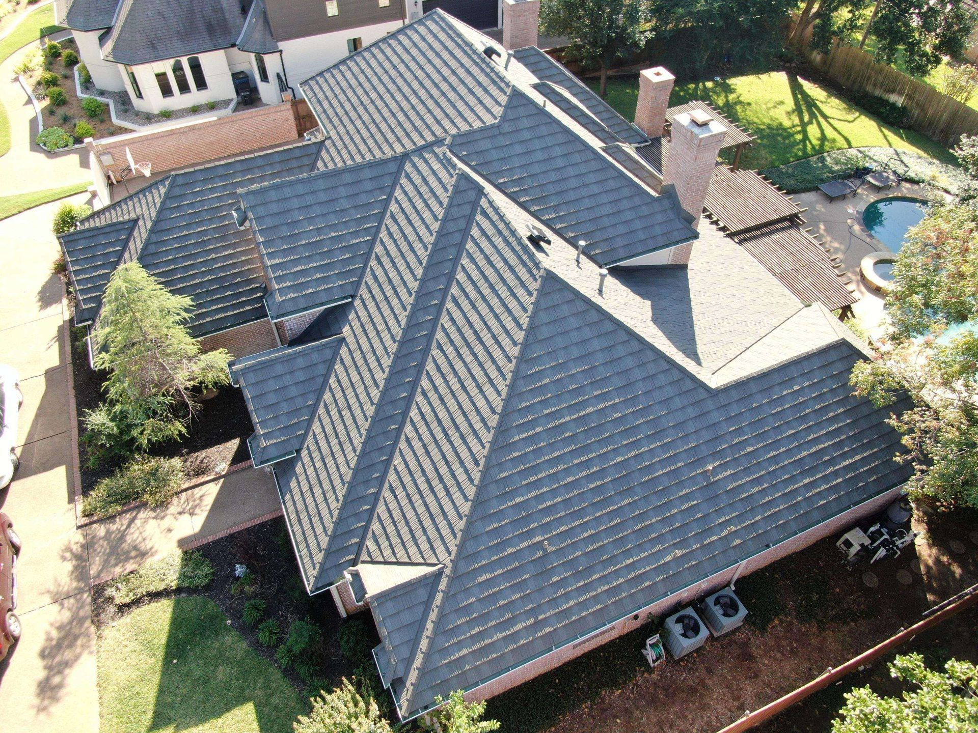 An aerial view of a dark shingled roof on a house with brick chimneys, surrounded by trees and a pool area.