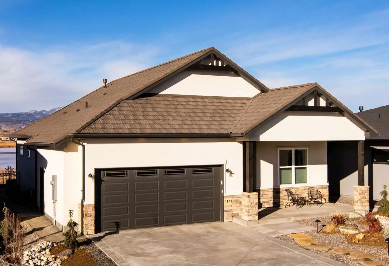 A beige single-story house with a dark brown garage, stone accents, a covered front porch, and a brown shingled roof.