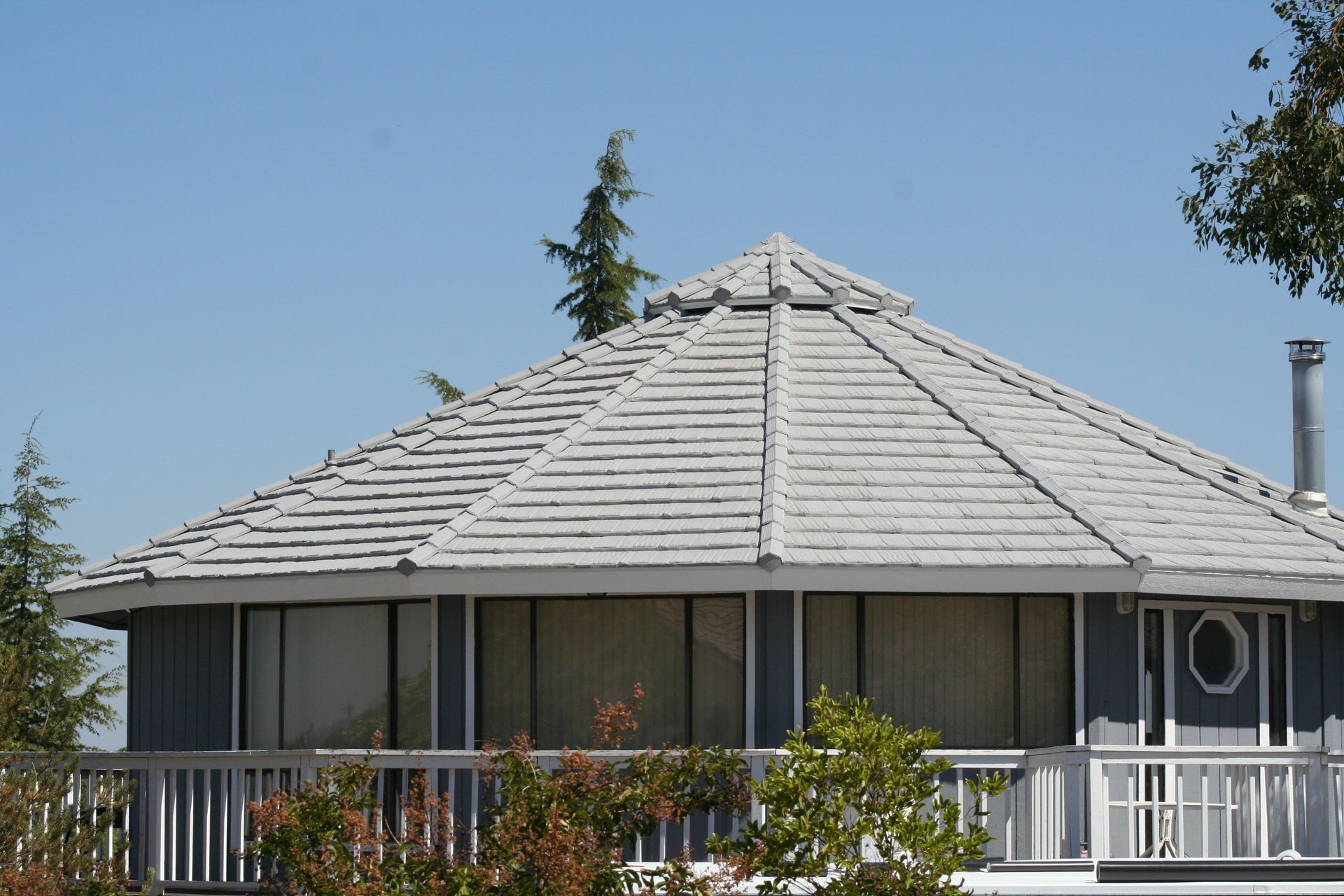 A blue, circular-shaped house with a grey, multi-faceted roof and a white railed deck, surrounded by trees.