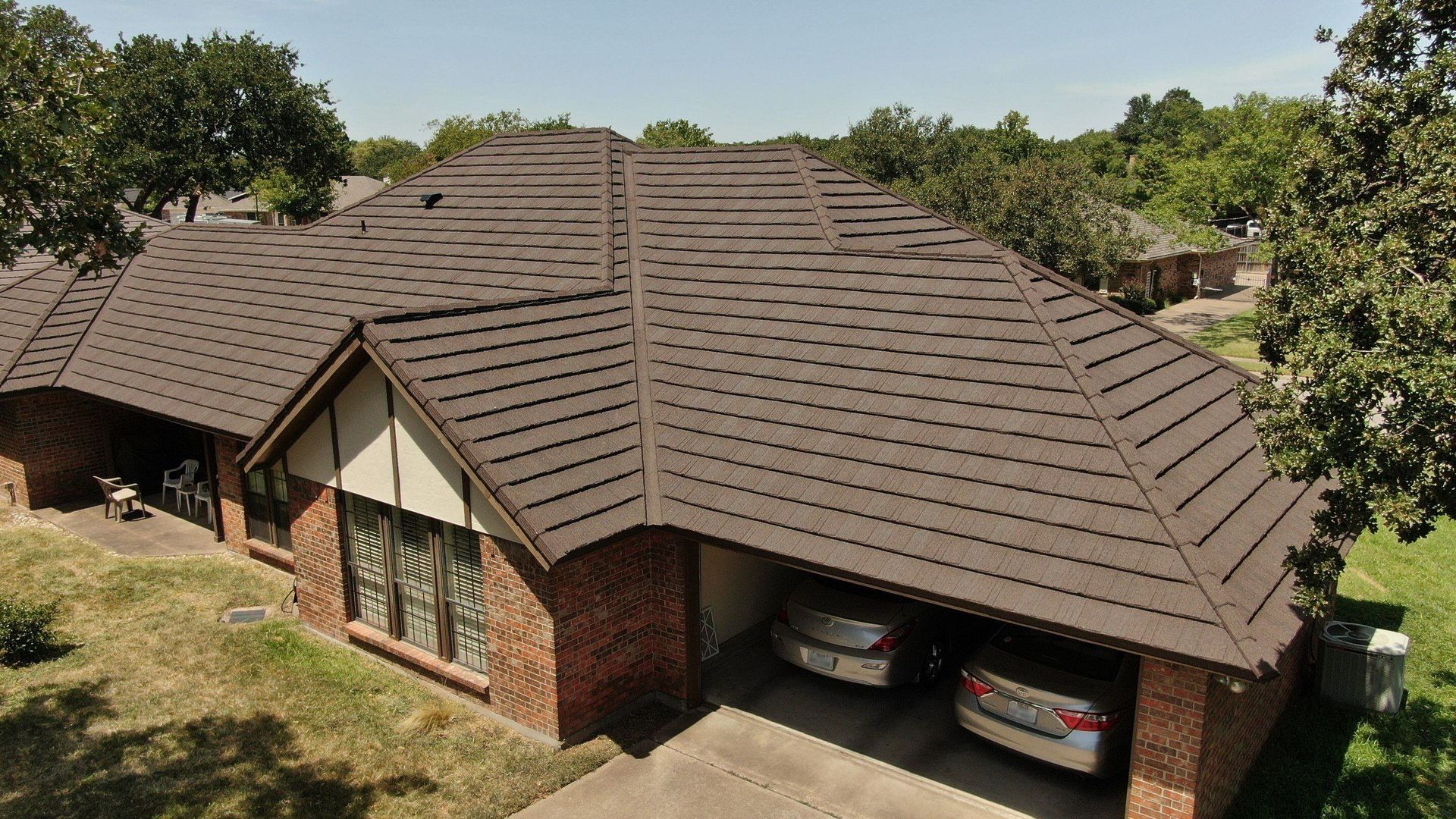 High-angle view of a brick house with a dark brown shingled roof, featuring a carport with two cars parked inside.