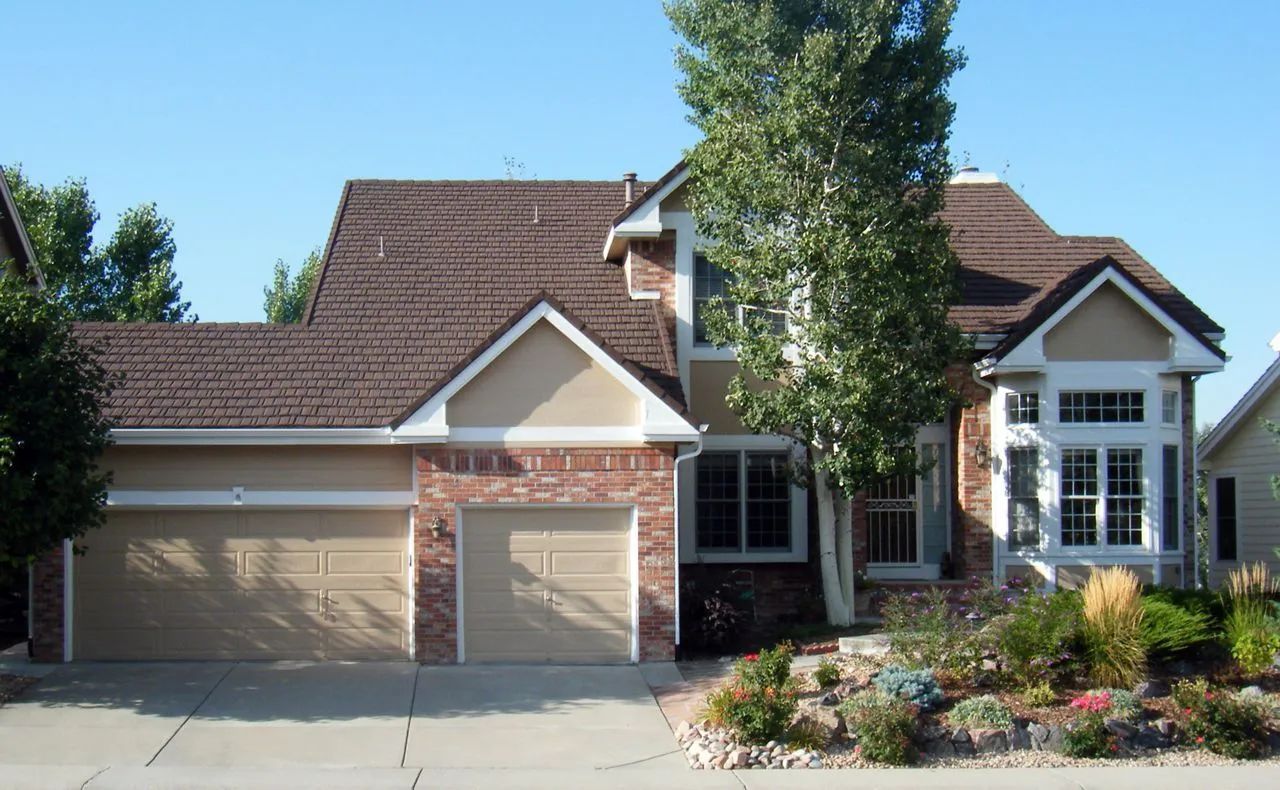 A two-story tan and brick suburban house with a multi-car garage, a peaked roof, and a landscaped front yard.