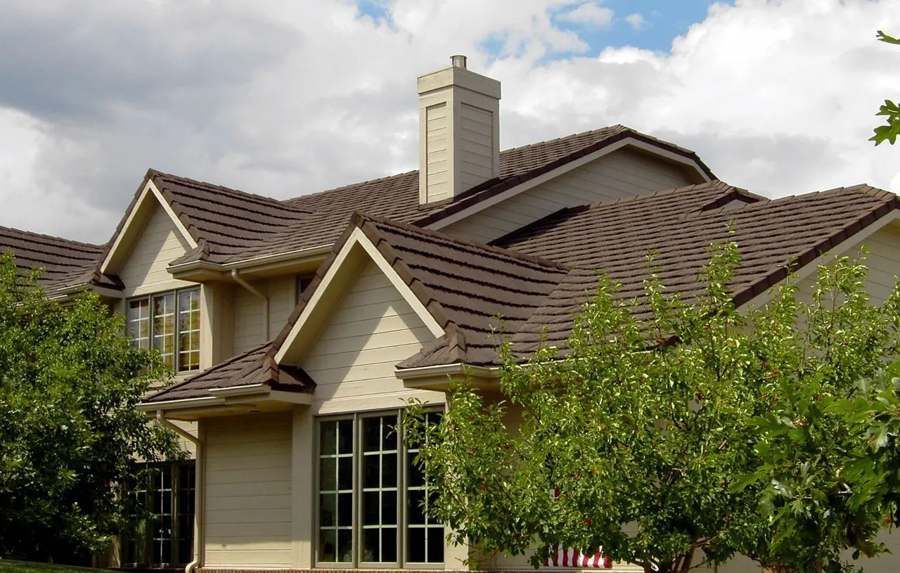 A multi-gabled house with tan siding and a dark brown tile roof, partially obscured by green trees under a cloudy sky.