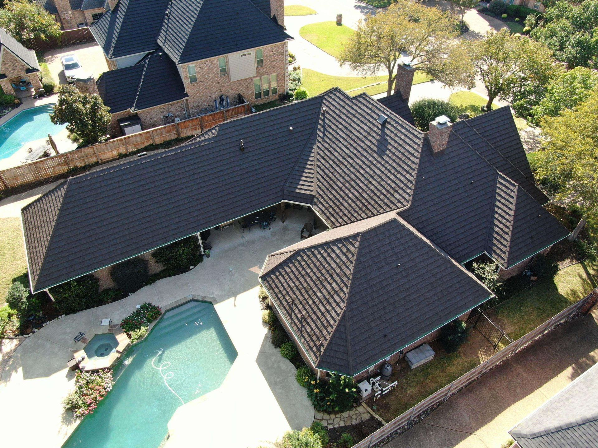 An aerial view of a residential house with a dark, shingled roof, a swimming pool, and surrounding green landscaping.