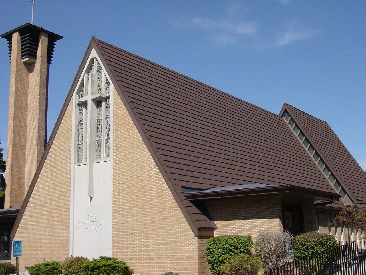 A tan brick church with a tall steeple, a prominent front cross window, and a dark brown gabled roof under a blue sky.