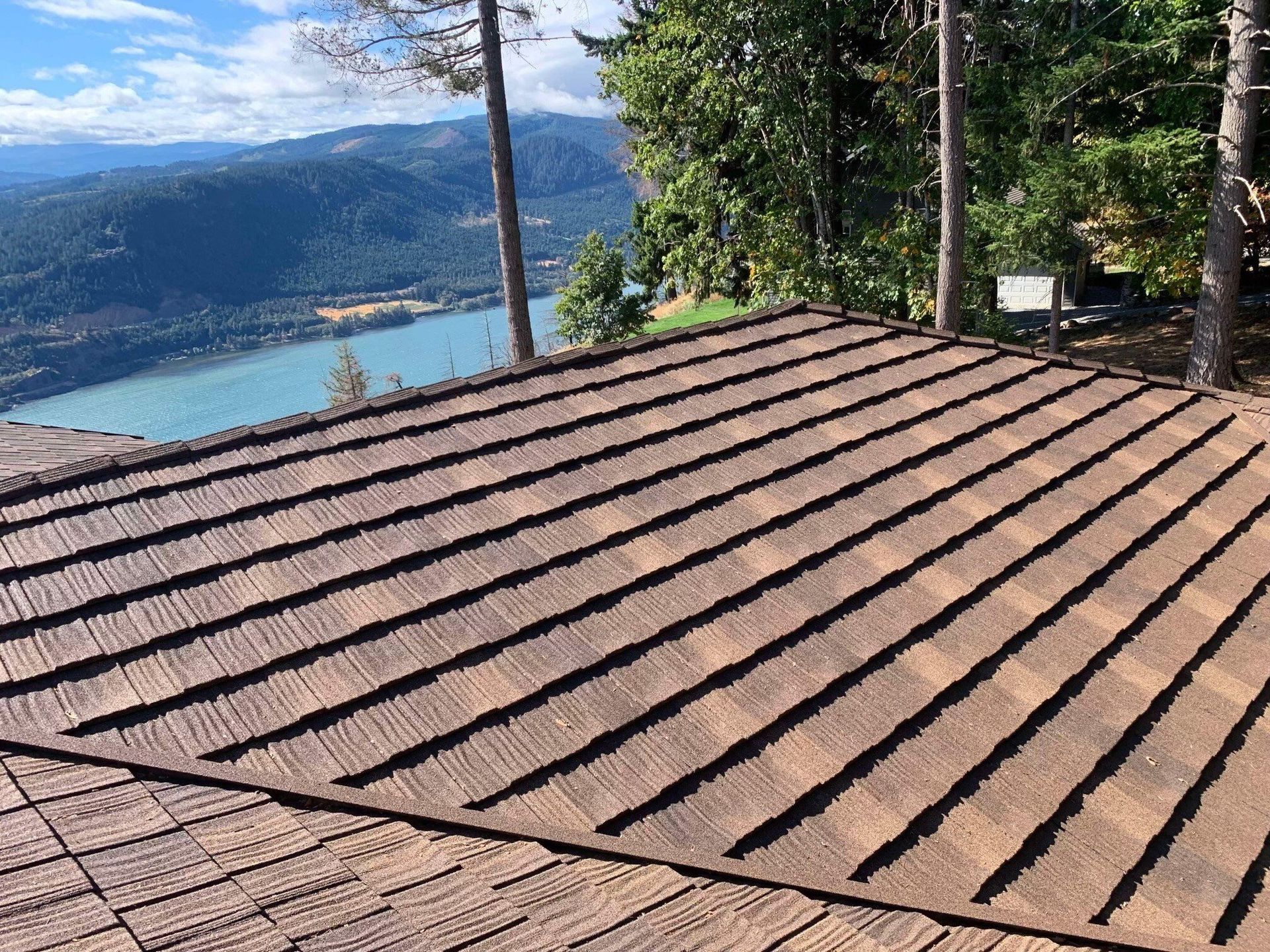 A view of a brown shingled roof overlooking a blue river valley, mountains, and pine trees.