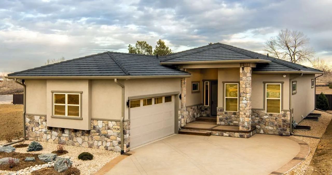 A single-story tan house with a stone foundation, gray shingled roof, two-car garage, and a front porch.
