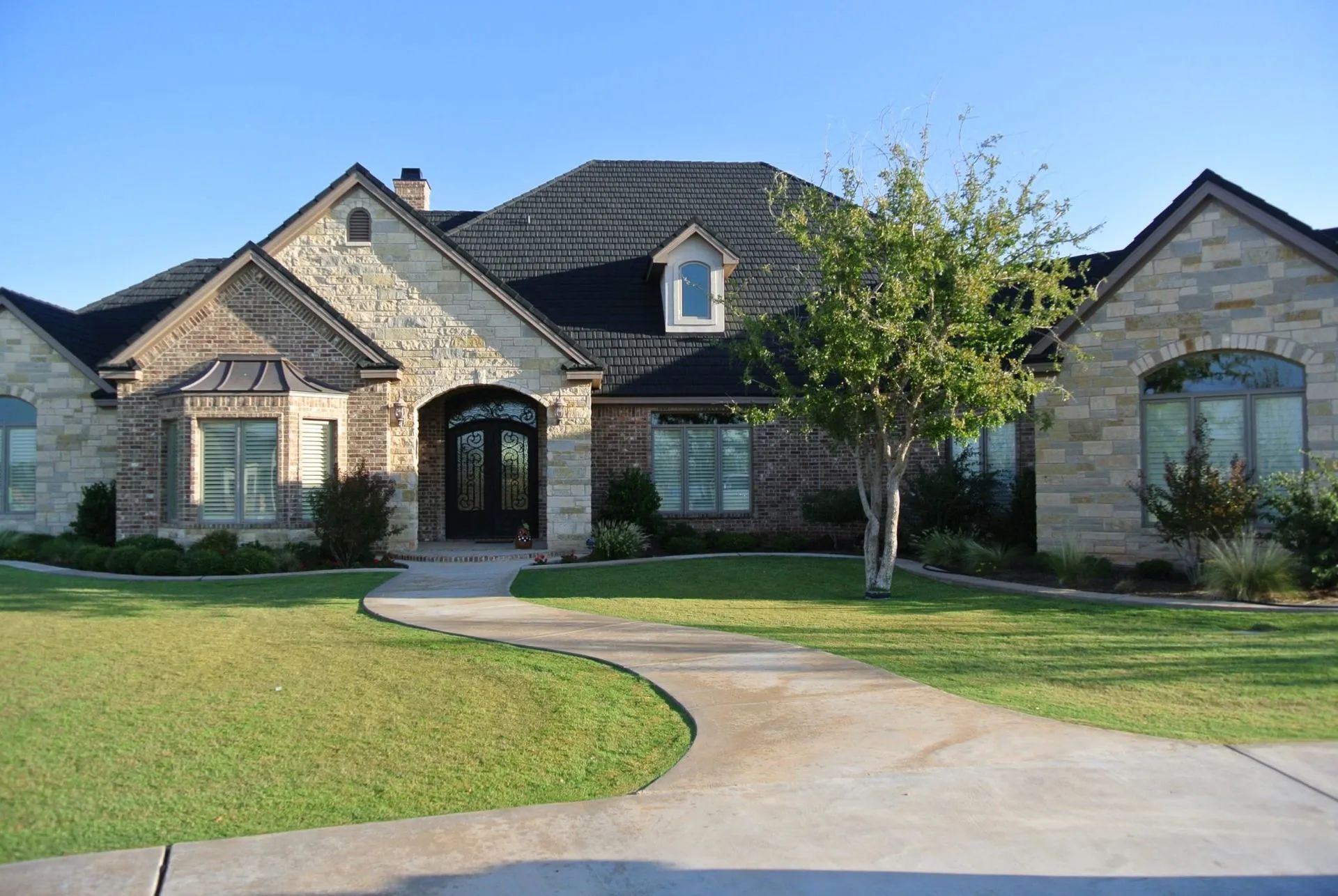 A large, single-story stone house with a dark gabled roof, a curved concrete walkway, and a small tree on a grassy lawn.