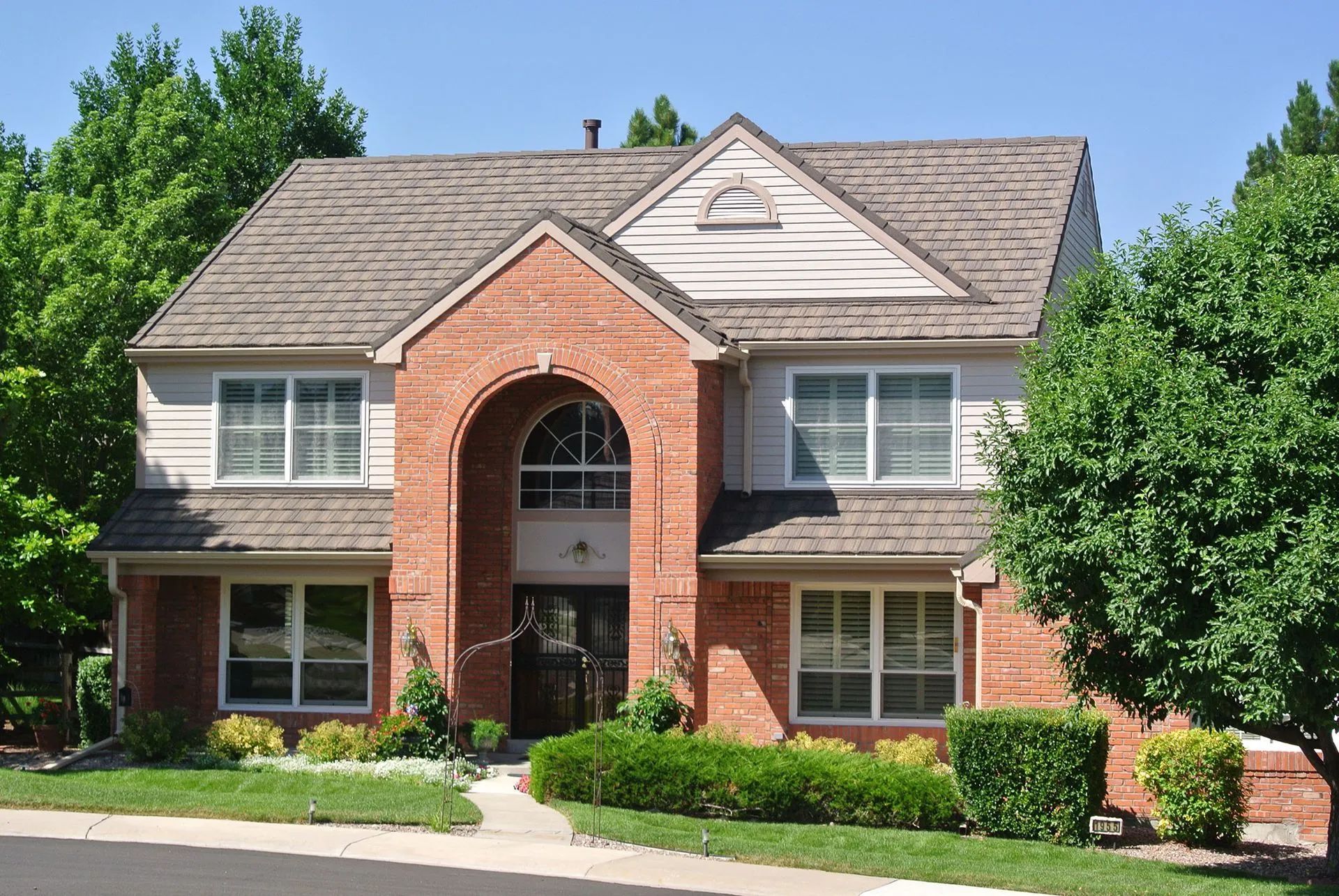 Two-story brick house with a front-facing gable, arched entryway, and gray shingled roof, surrounded by green landscaping.