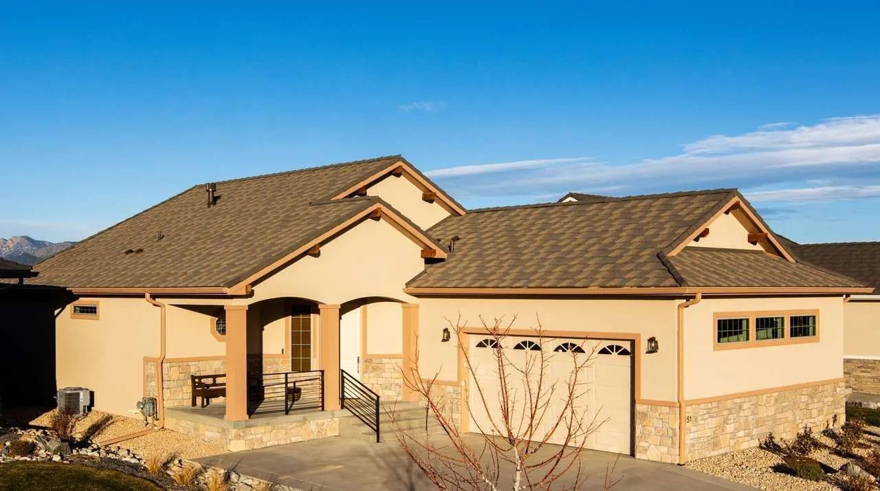 A single-story tan house with a stone base, a covered front patio, and an attached two-car garage under a blue sky.
